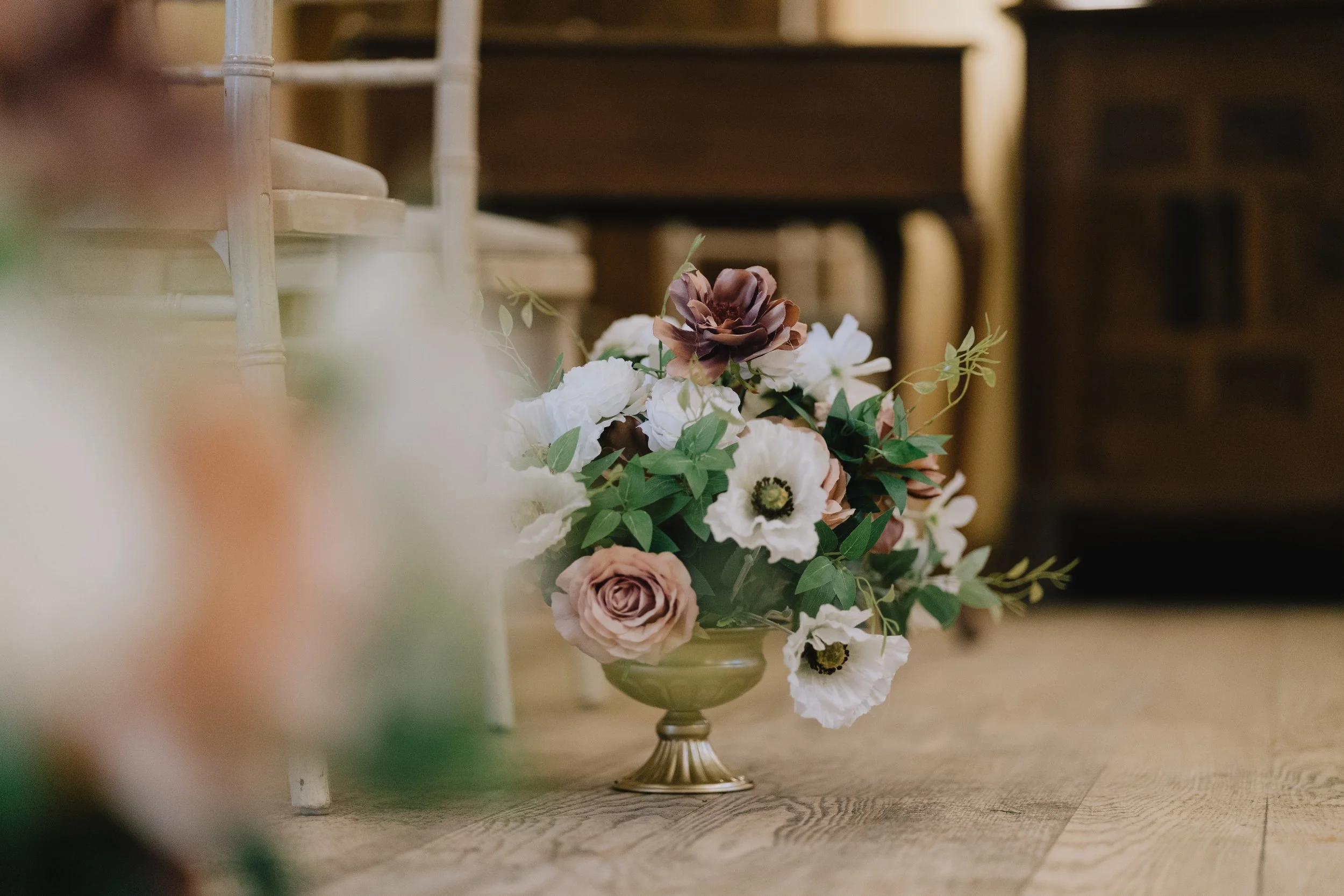A floral arrangement in a decorative brass vase with white, pink, and dark purple flowers, placed on a wooden floor in a cozy room.