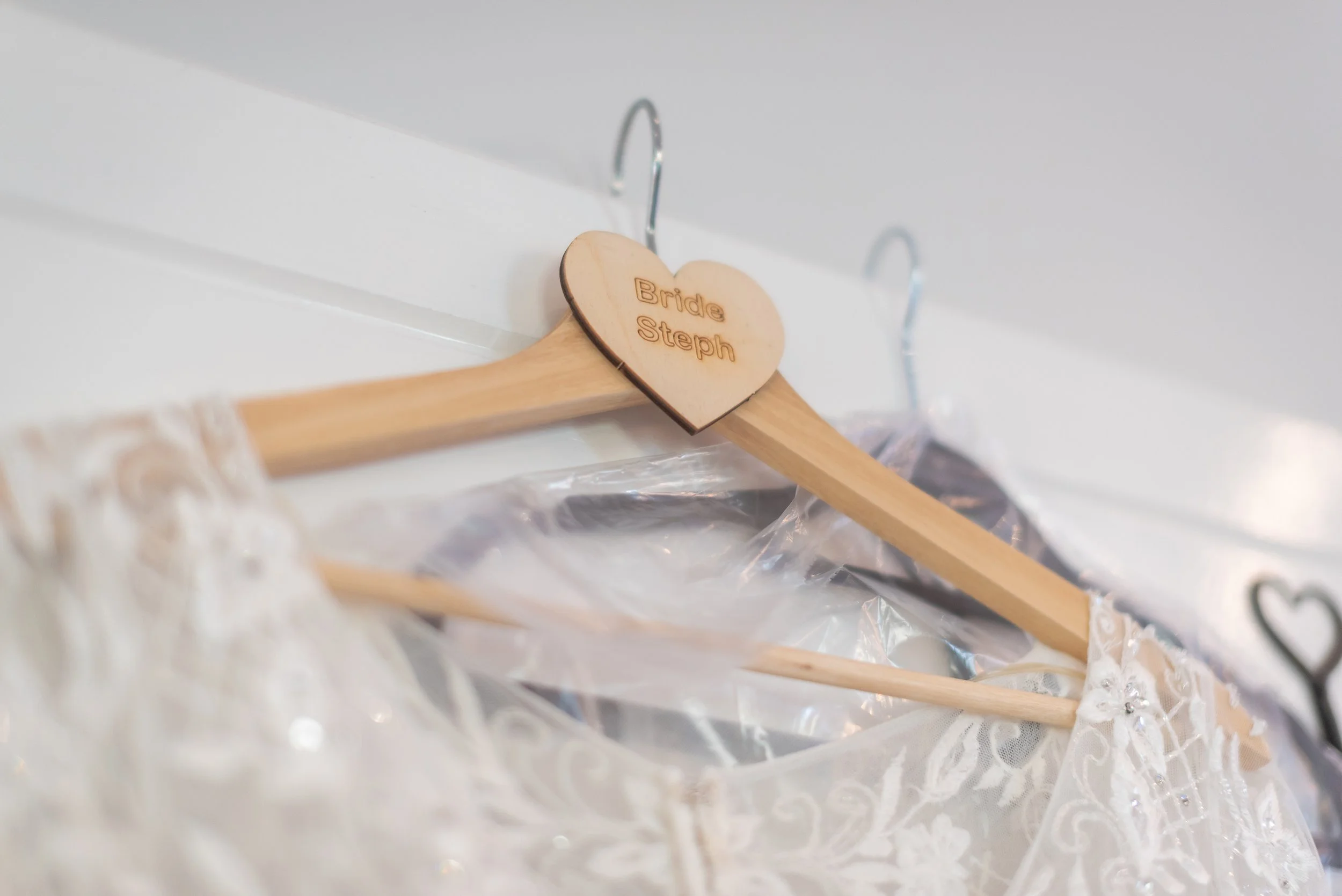 A wooden hanger with a heart-shaped top engraved with 'Bride Steph' holding a wedding dress with lace details and embellishments taken by Midlands Wedding Photographer.
