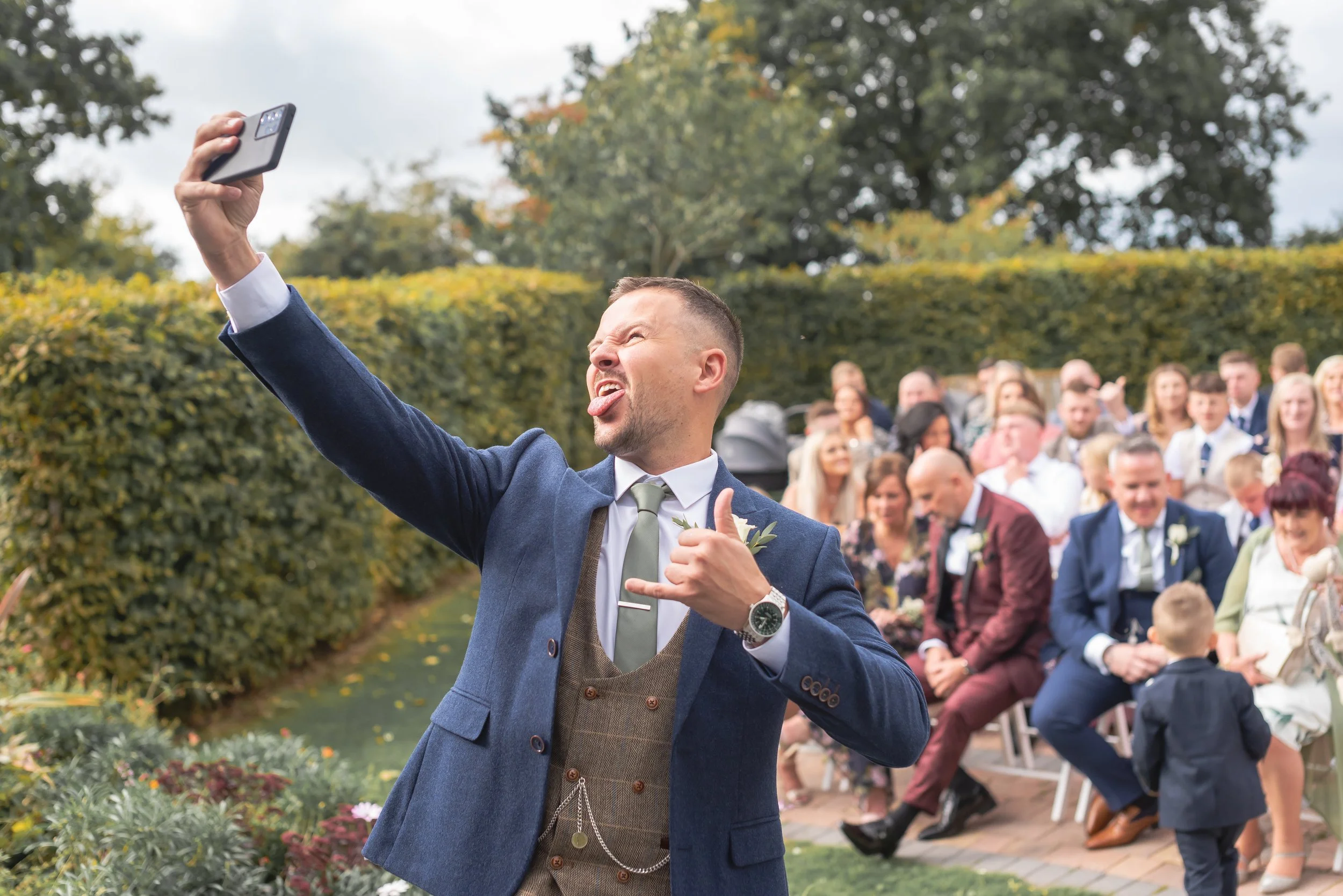 Man in a blue suit taking a selfie at an outdoor wedding ceremony, with guests seated in the background at Wootton Park in Midlands