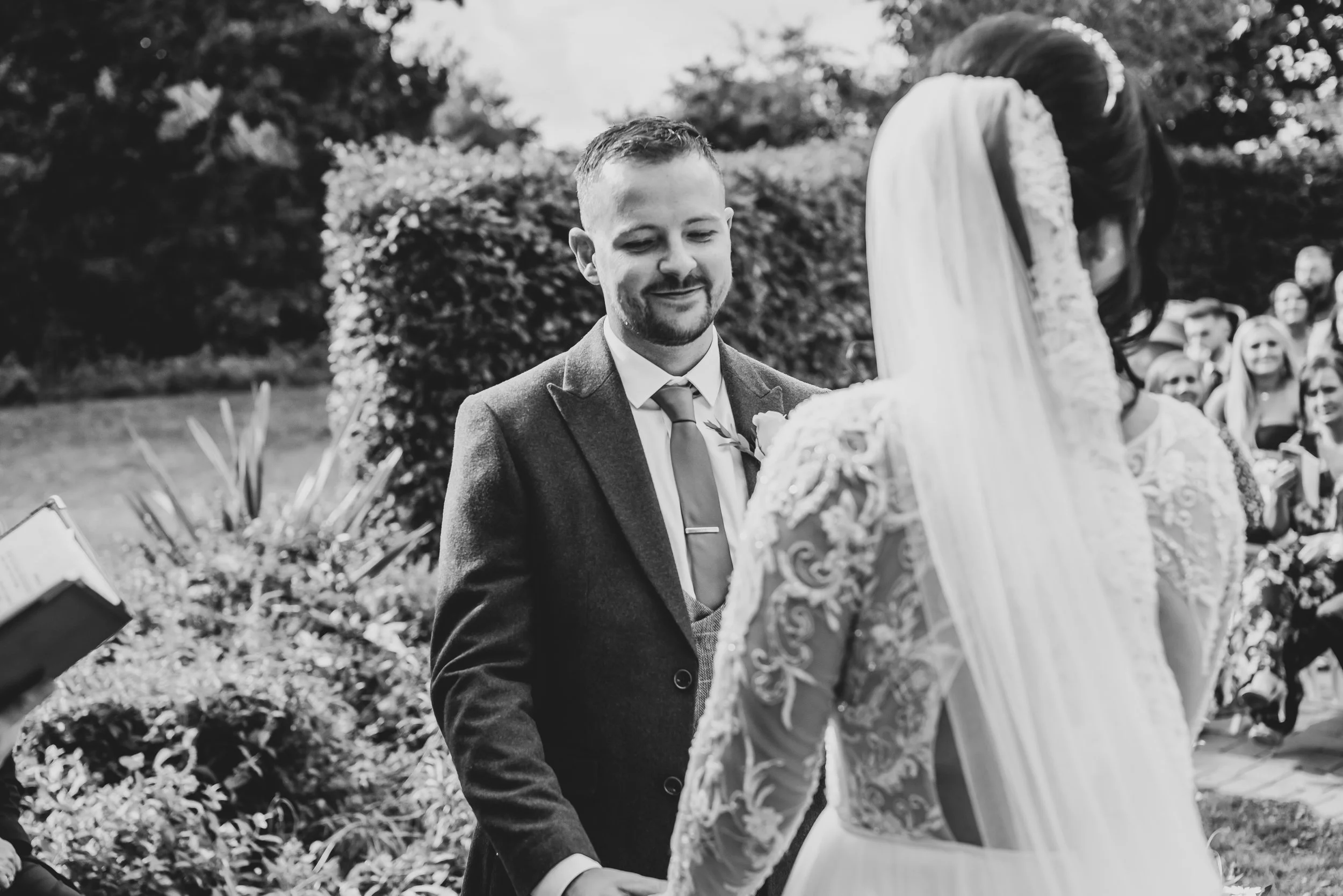 A black and white photo of a bride and groom exchanging vows outdoors during their wedding ceremony, with guests in the background at Wootton Park in Midlands