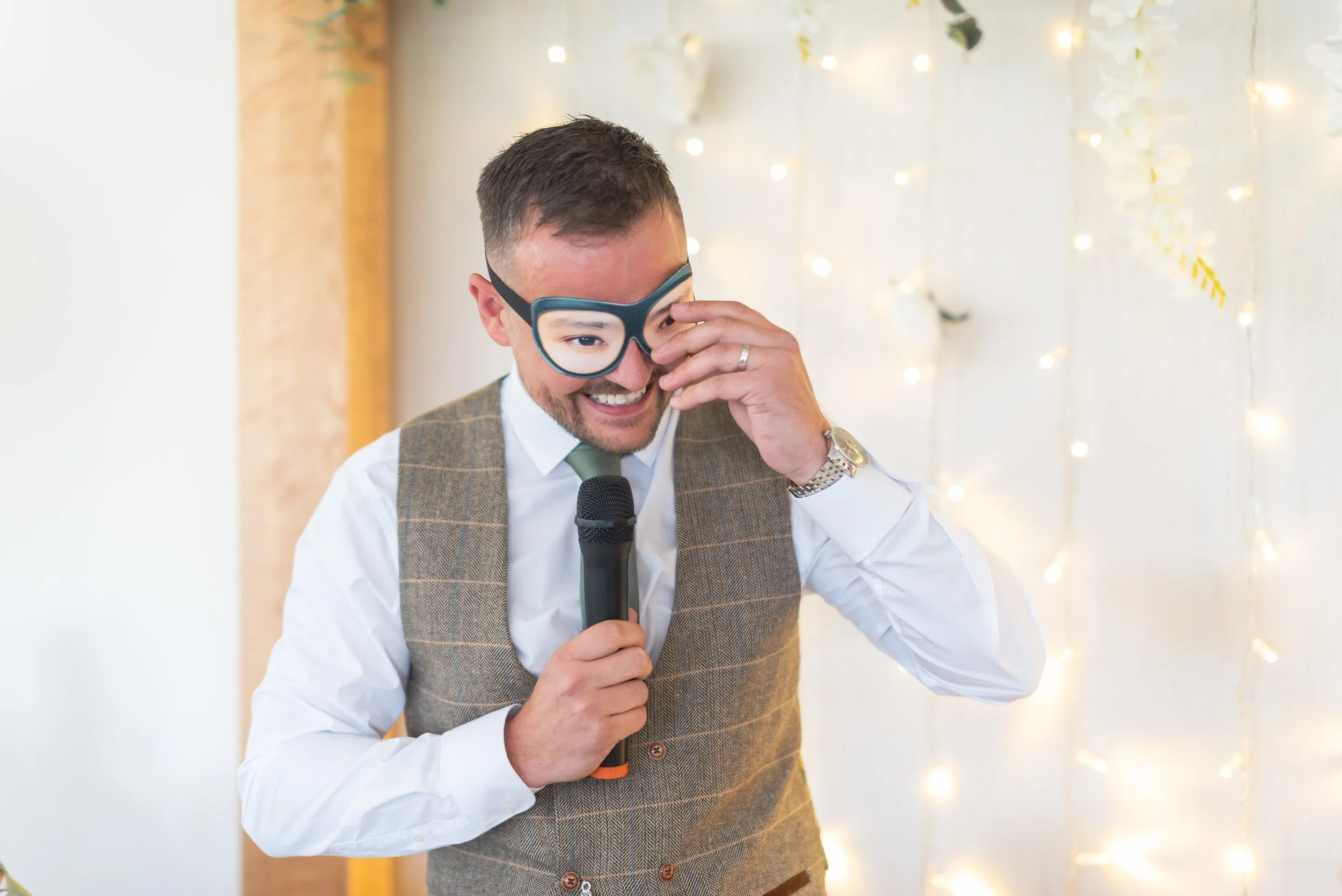 Wootton Park photographer captures groom wearing glasses, a vest, and a white shirt is holding a microphone and smiling. He is adjusting his glasses with one hand during his speech.