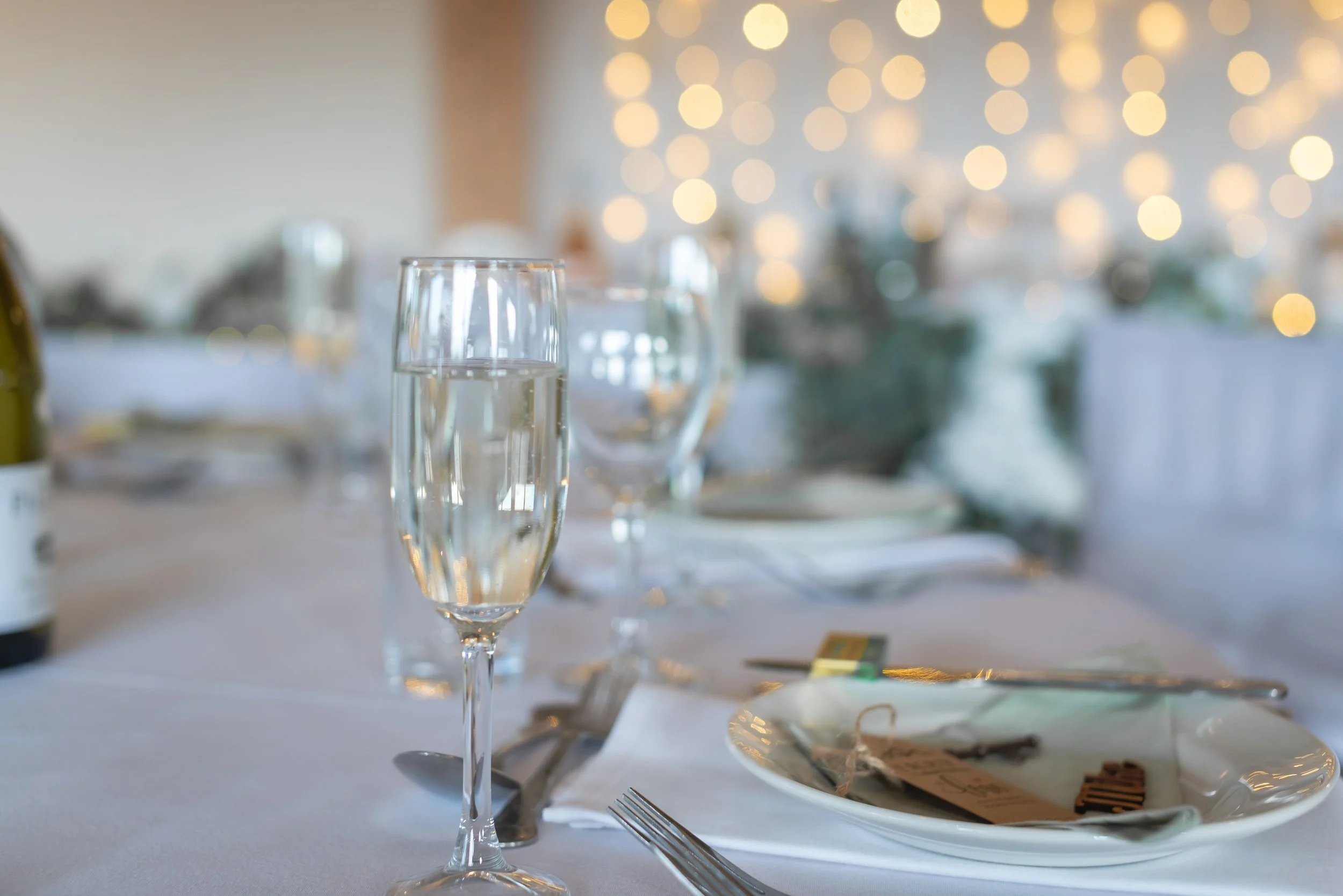 A close-up of a table setting at a wedding or event, featuring a champagne flute, wine glass, plate, utensils, and napkin, with blurred decorative lights in the background at Wootton Park in Midlands
