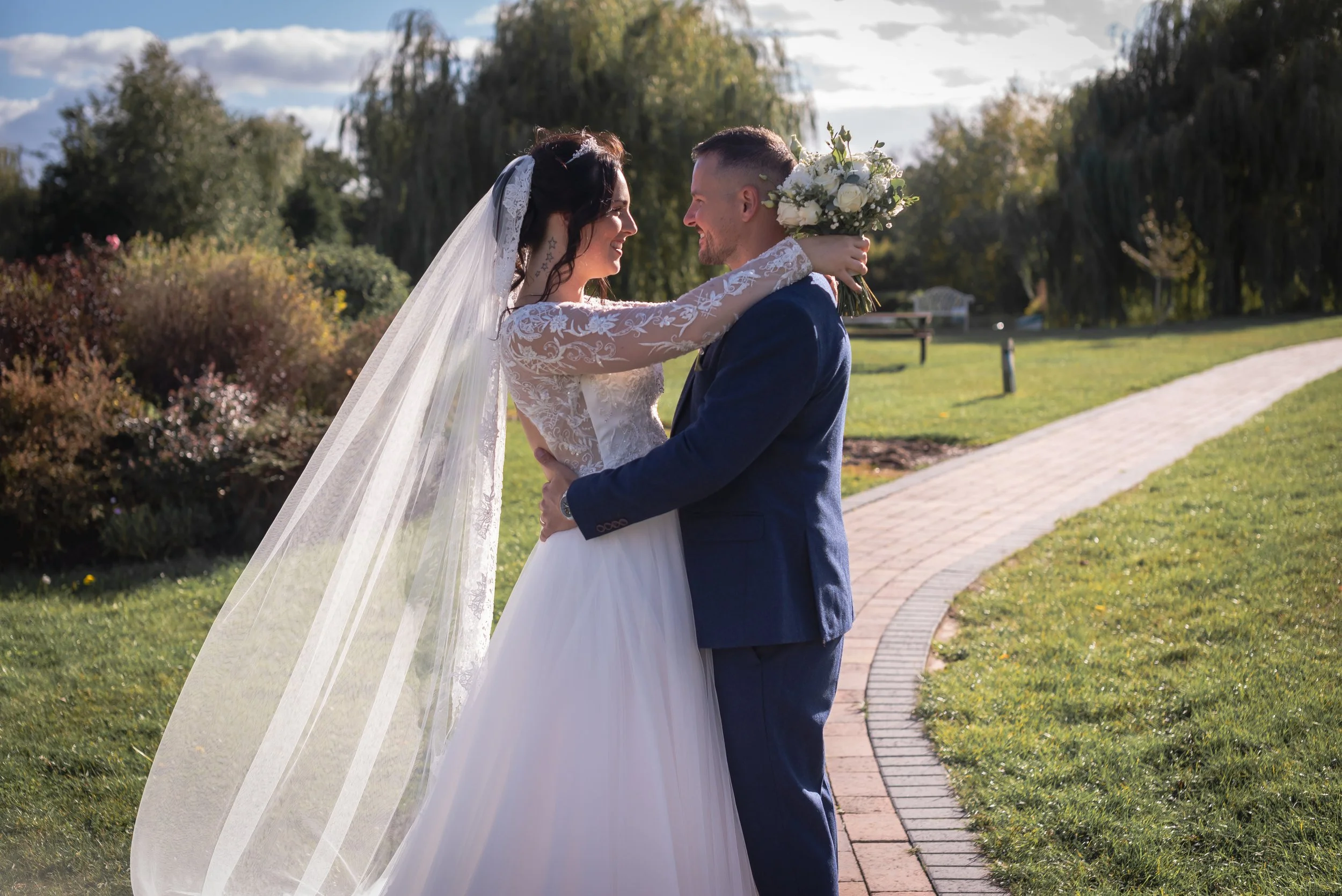 A bride and groom embrace in a park during their wedding celebration. The bride wears a lace wedding dress with a long veil, and the groom wears a dark blue suit. The bride holds a bouquet of white flowers, and they smile at eachother at Wootton Park