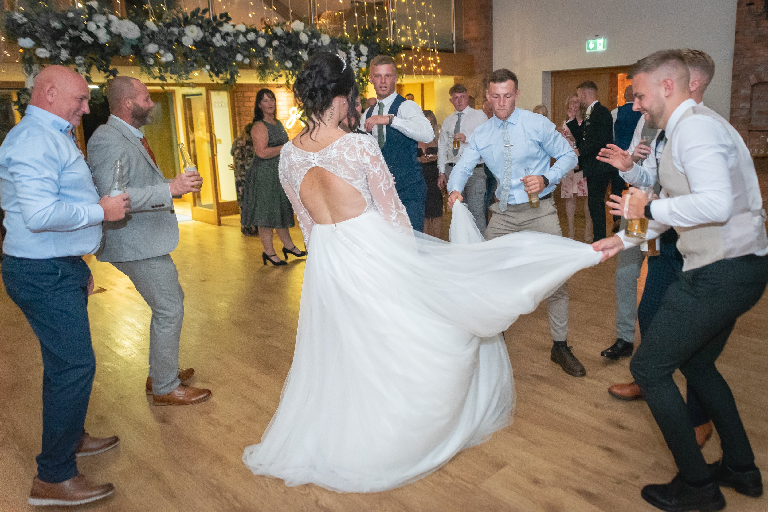 A bride in a white wedding gown dancing with guests at a wedding reception. The guests are holding drinks and dancing around her on a wooden floor, with wedding decorations and fairy lights in the background at Wootton Park in Midlands