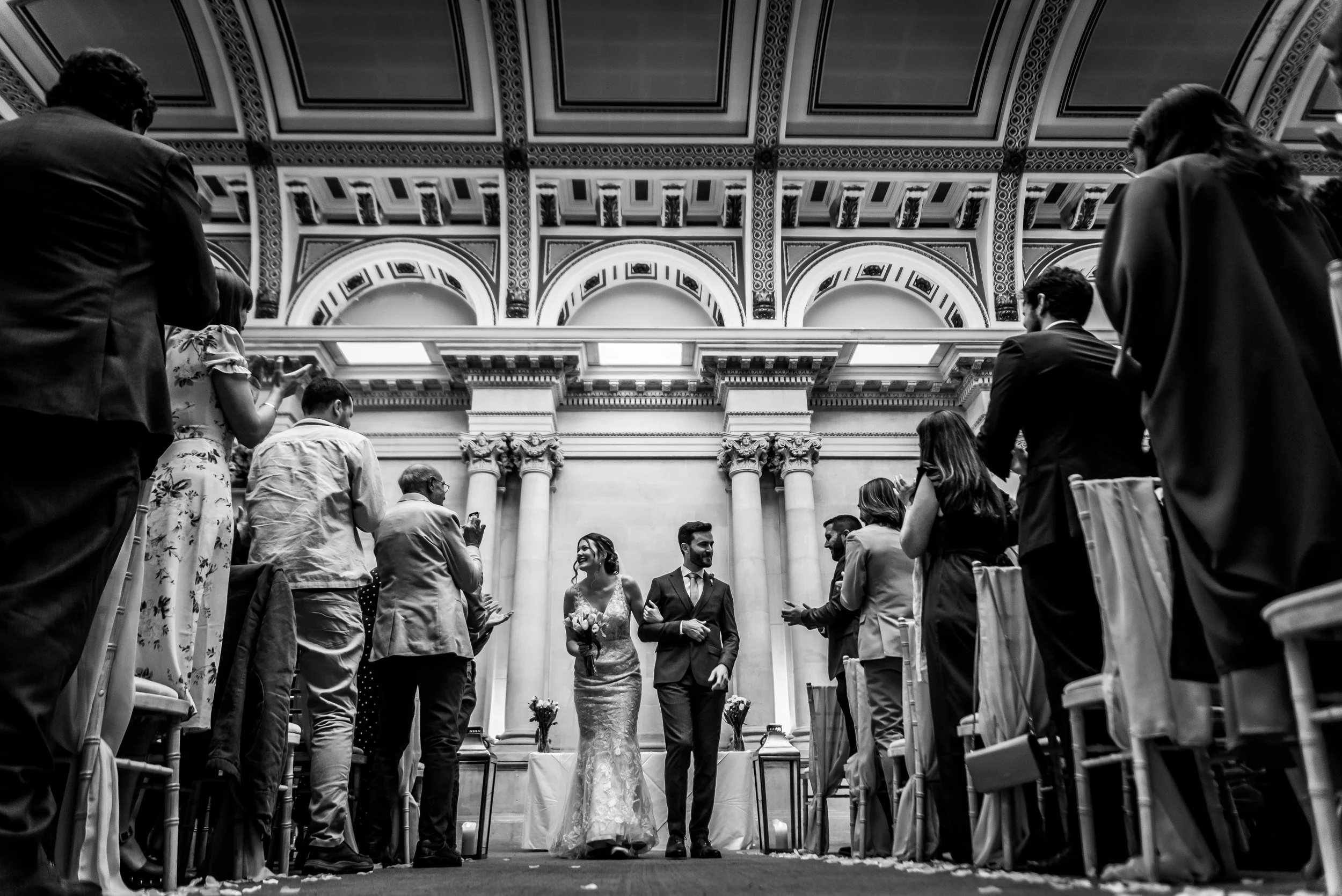 Black and white photo of a couple walking down the aisle during a wedding ceremony in a grand hall with ornate architectural details at Bristol Harbour Hotel taken by Bristol Wedding Photographer