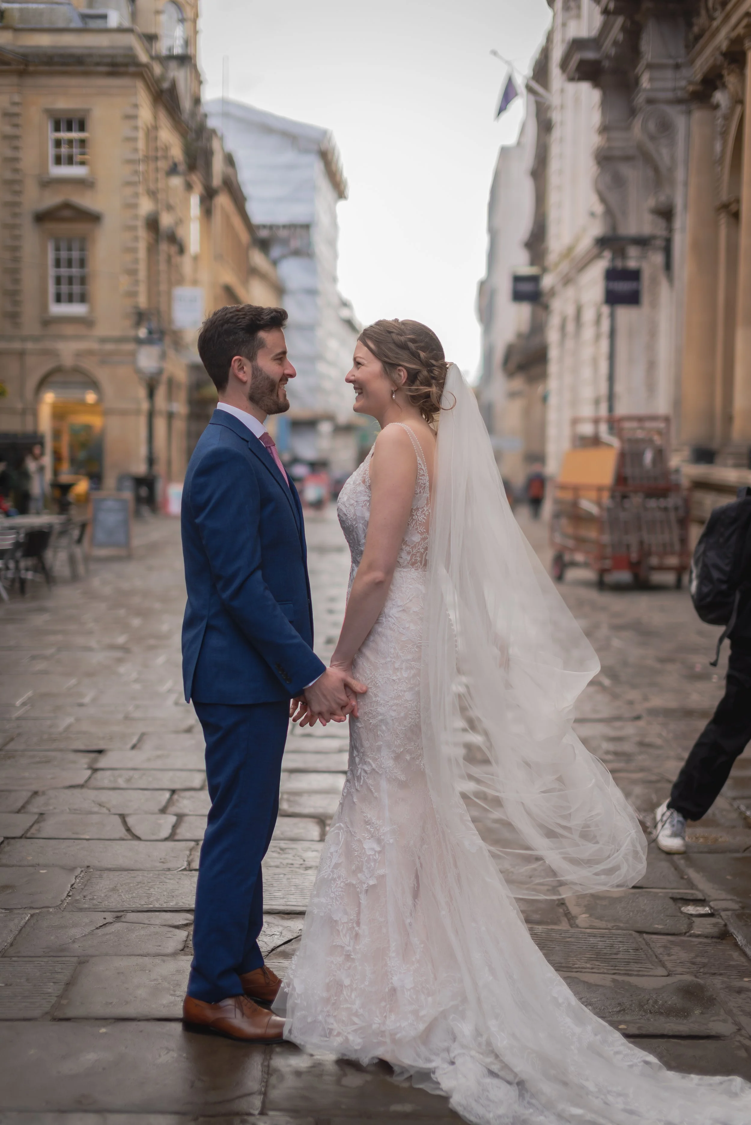 A bride and groom holding hands and smiling at each other on a city street with historic buildings on Corn Street in Bristol.