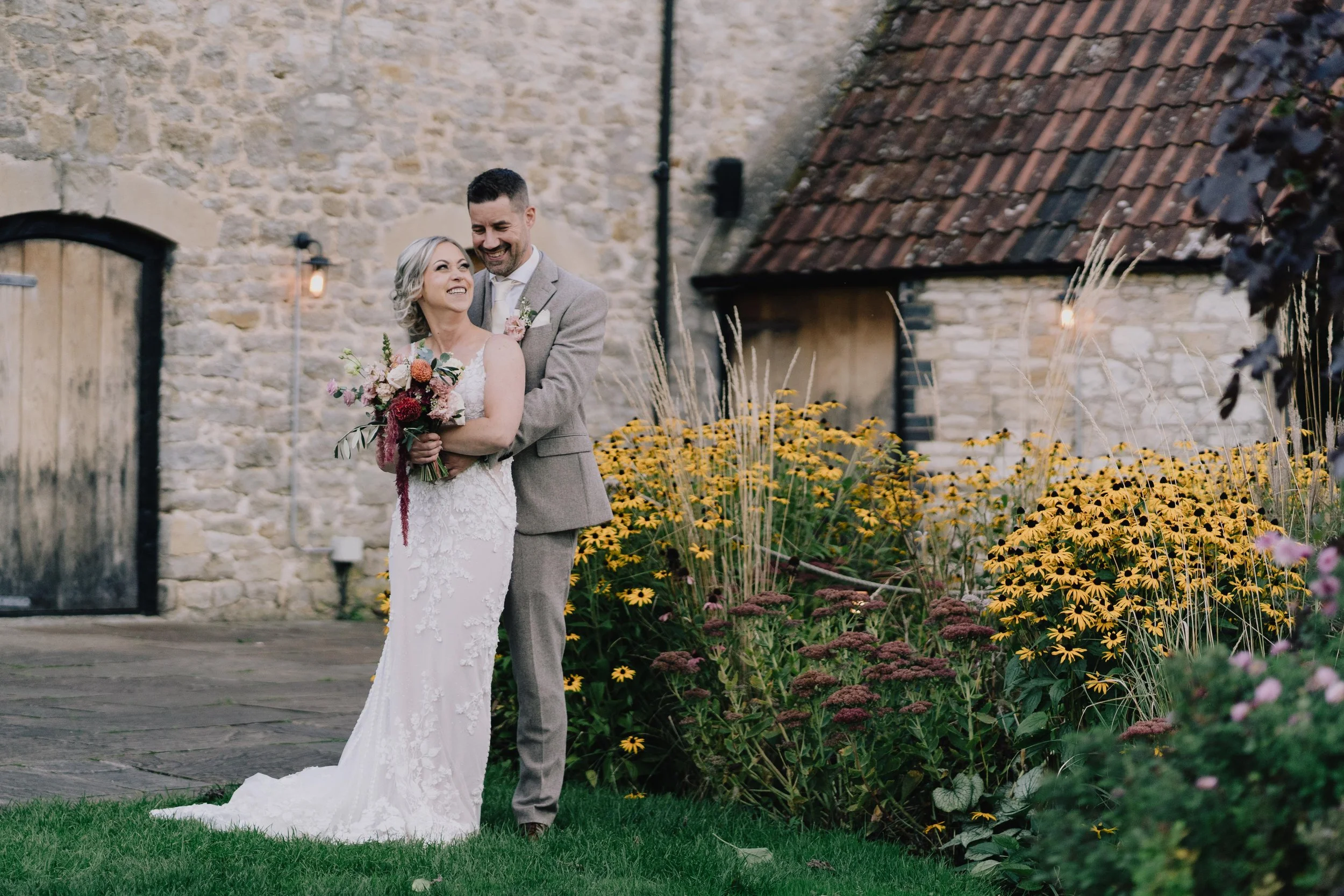 Bride and groom standing together outdoors, smiling, with historic stone building and yellow flowers in background at Priston Mill taken by Bristol Wedding Photographer
