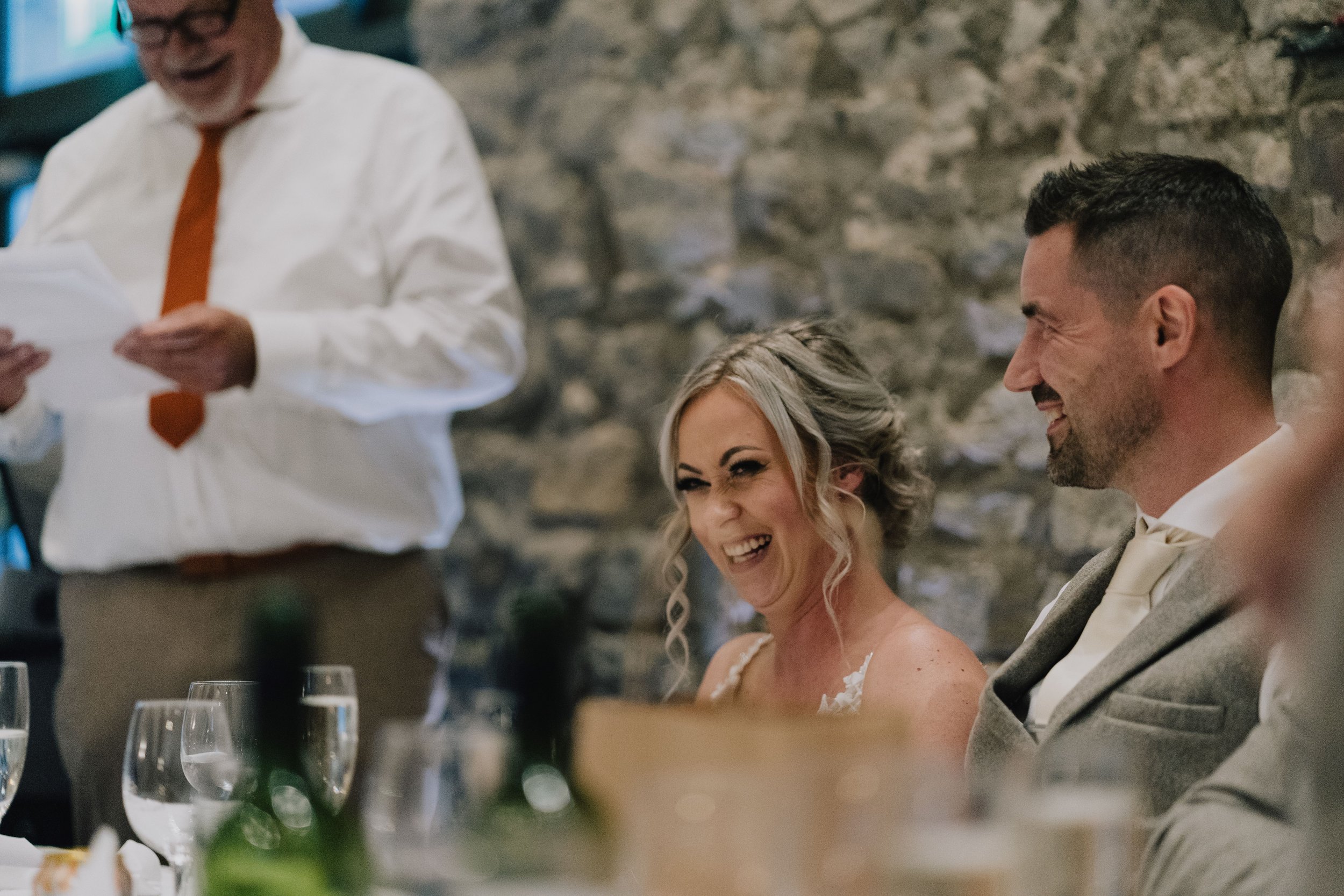 A woman with blonde hair styled in curls and a man with short dark hair and a beard, both smiling and laughing, sitting at a table during a celebration or wedding reception at Priston Mill