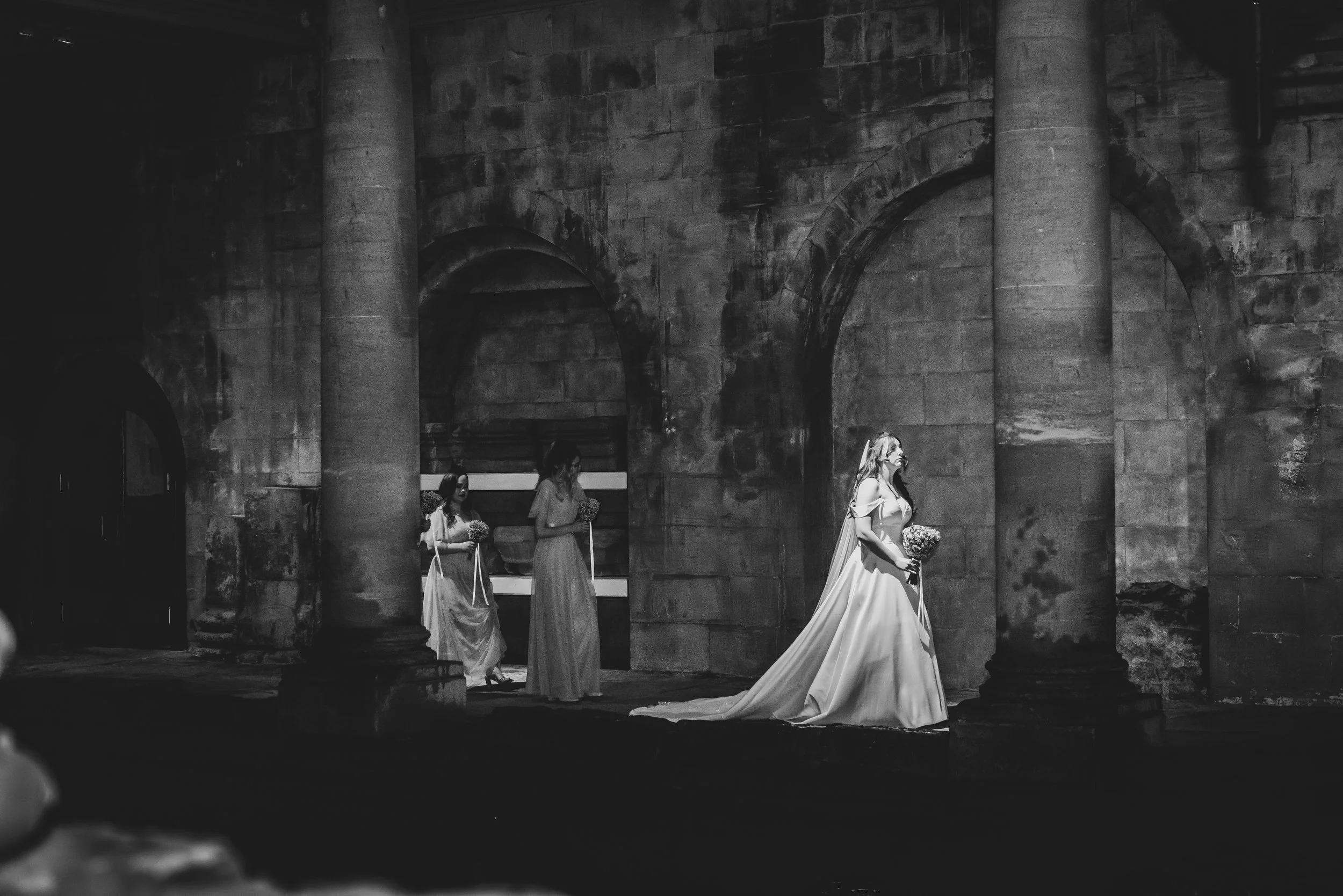 A black and white photo of a woman in a wedding gown holding a bouquet, walking outside near large stone columns, with two other women in dresses following her at Roman Baths