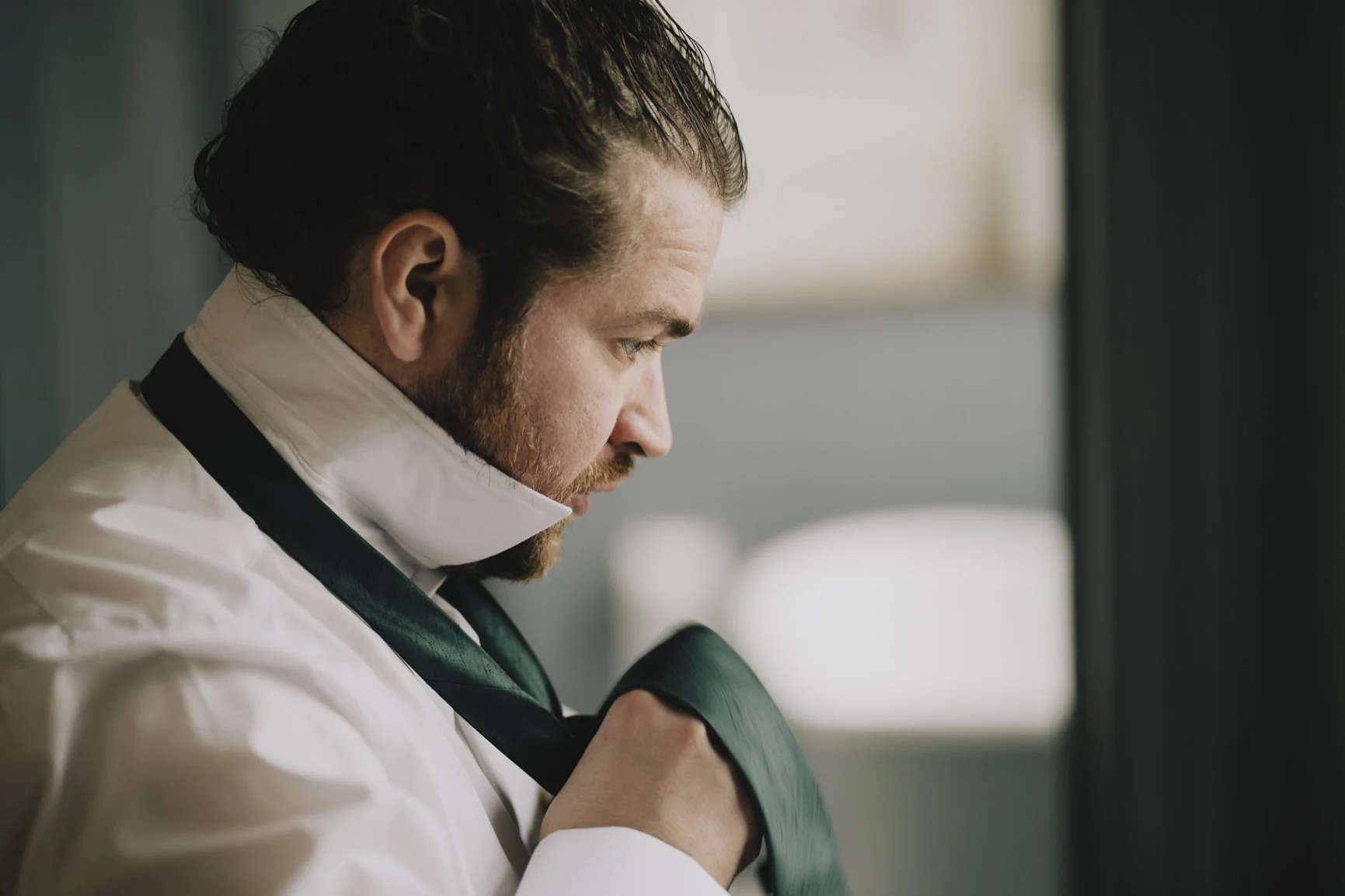 Grrom with dark hair and a beard, dressed in white medical attire, adjusting a dark tie in front of a window ready to have his wedding captured by Greenhill Wedding Photography