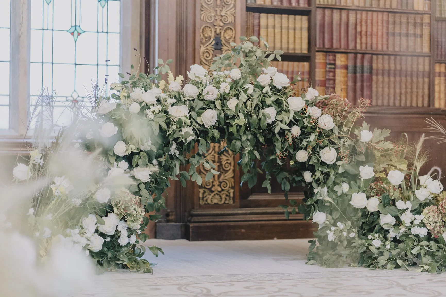 A floral arch by Blooms at Breach with white roses and greenery in front of a wooden wall with bookshelves and a stained glass window at Clevedon Hall