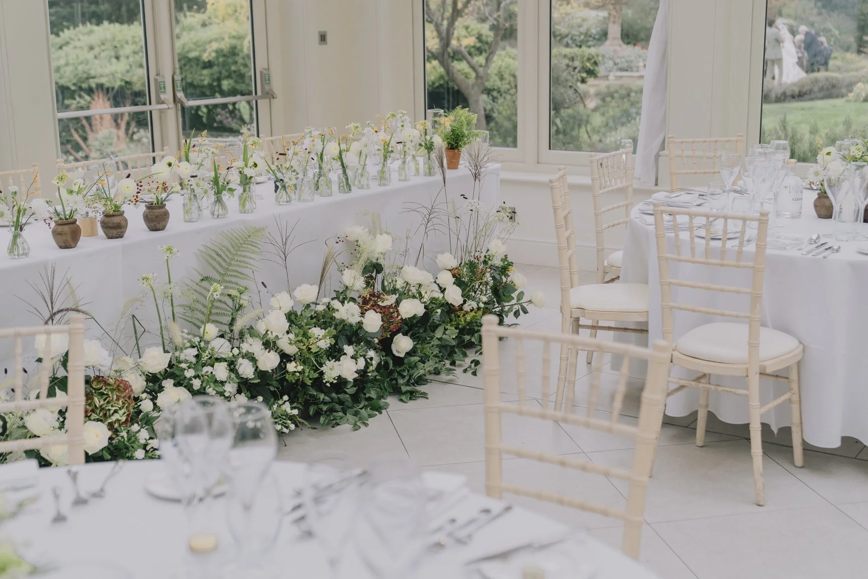 Wedding reception table decorated by Blooms at Breach with white flowers, glassware, and beige chairs, with window views of a garden and wedding guests outside.