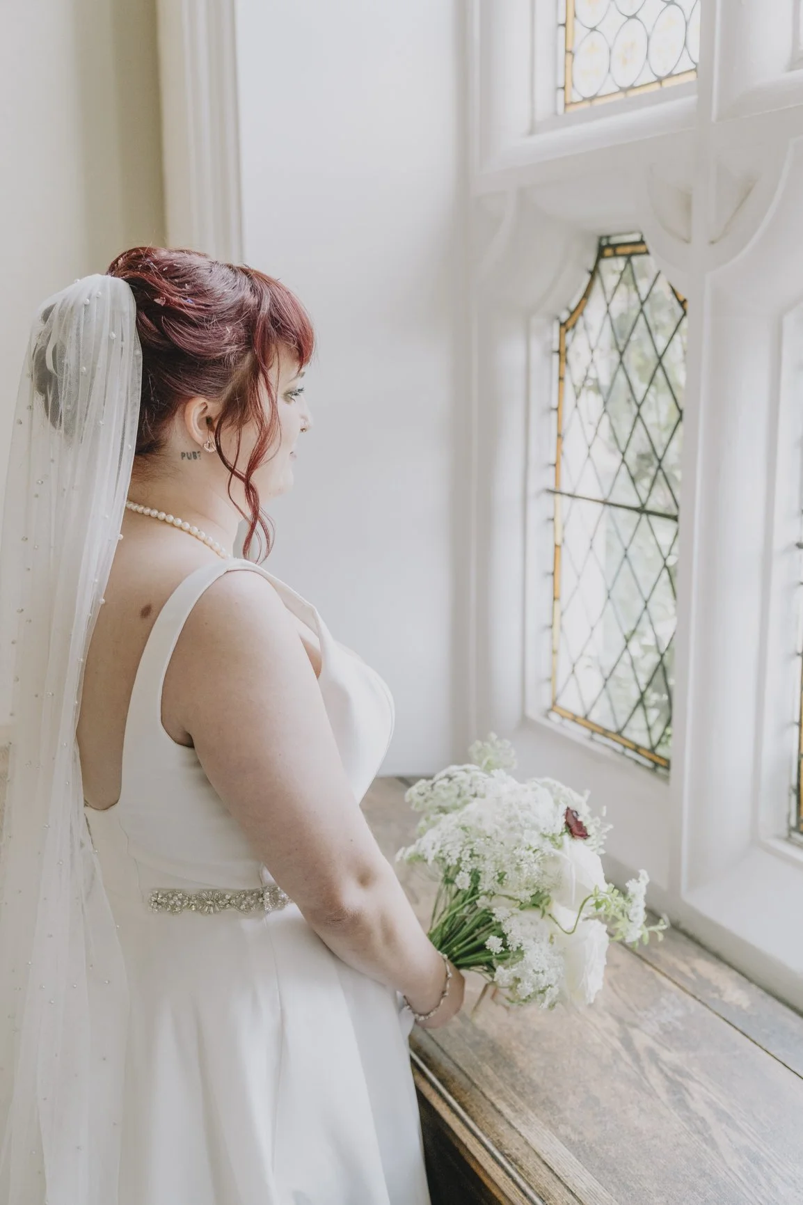 A bride with red hair wearing a white wedding dress holding a bouquet of white flowers and looking out a window taken by Clevedon Hall Wedding Photographer.