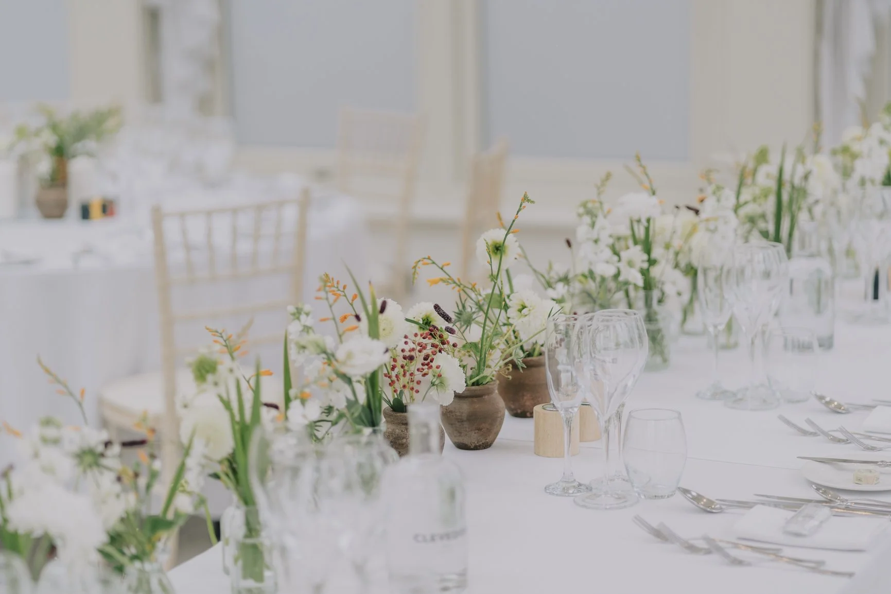 Elegant table setting with white flowers in small brown vases, empty wine glasses, and silverware on a white tablecloth at a bright, softly lit event space at Clevedon Hall.