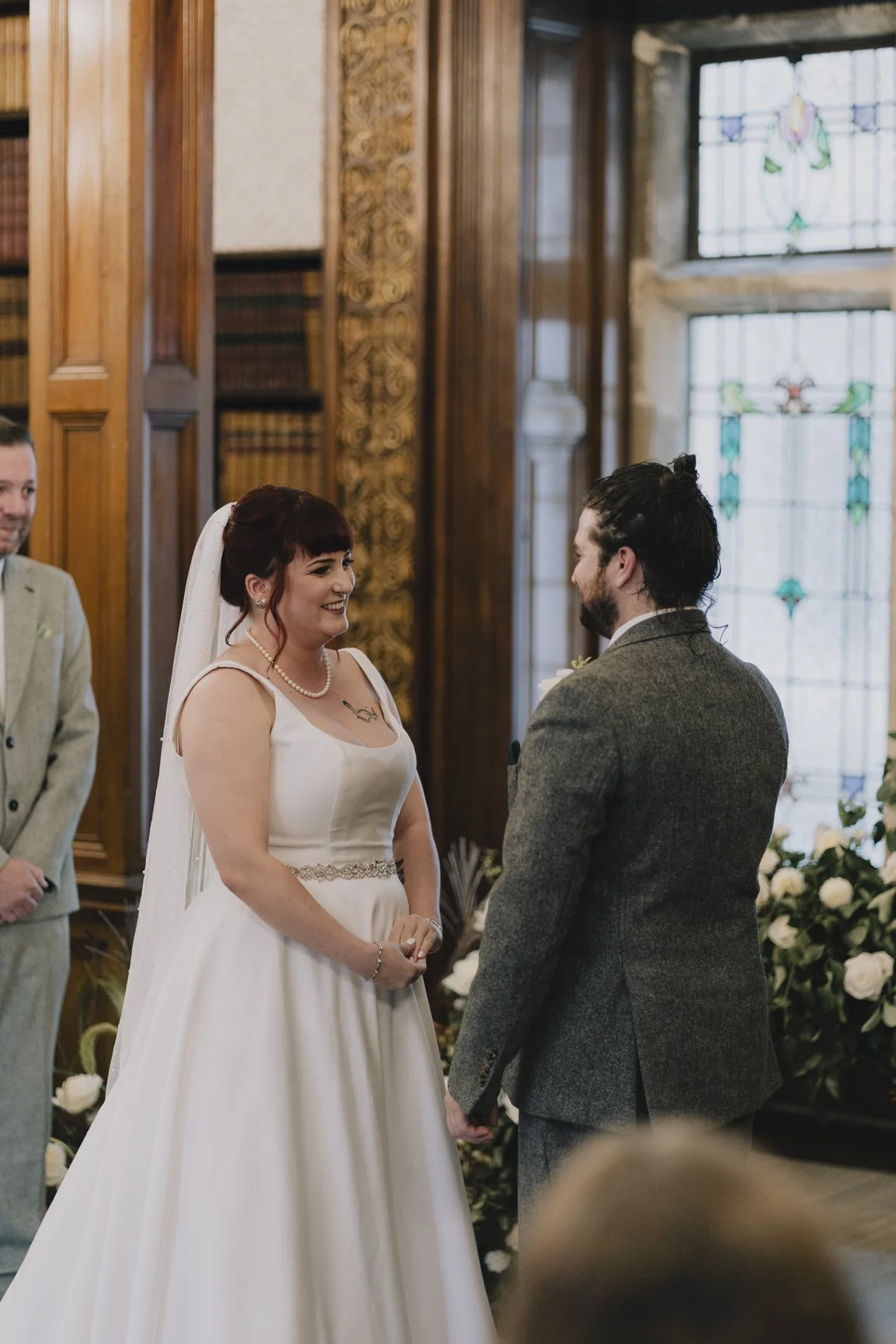 A bride and groom holding hands and smiling at each other during their wedding ceremony inside a decorated church with stained glass windows and floral arrangementsd at Clevedon Hall.