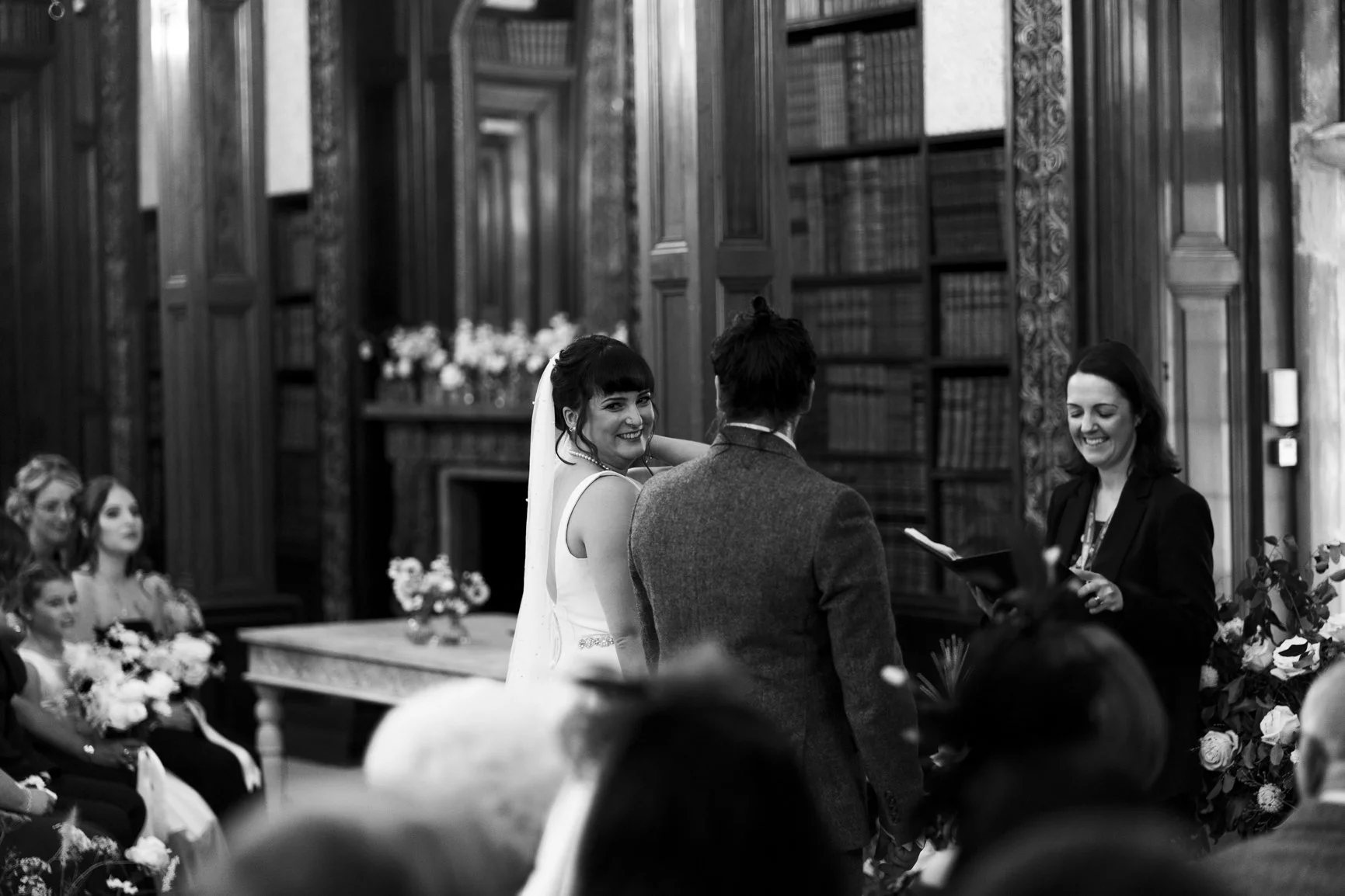 A black and white photo of a wedding ceremony with a bride and groom facing a female officiant, with seated guests in the background, in an ornate room with bookshelves and wooden paneling at Clevedon Hall taken by local wedding photographer.