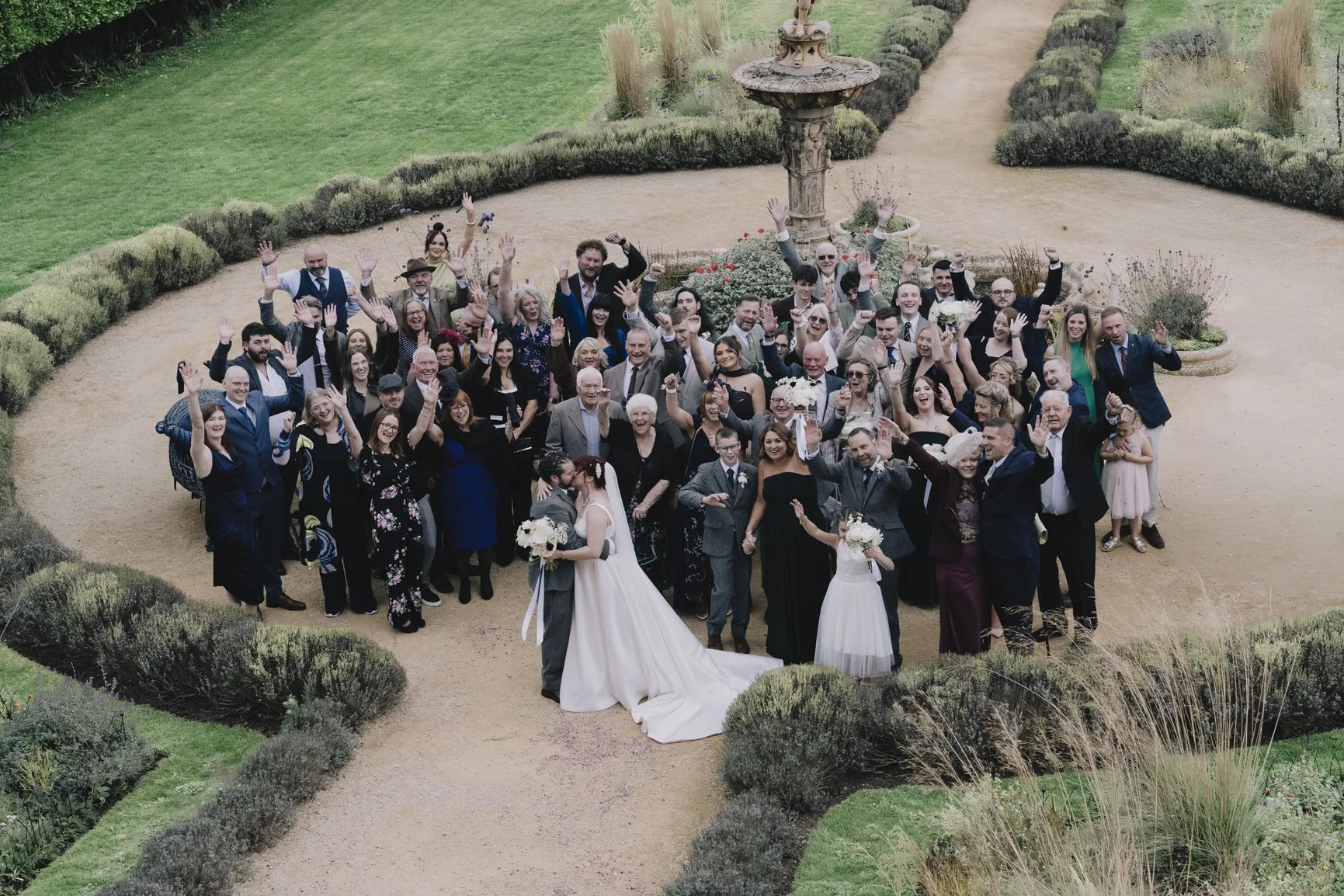 A large group of wedding guests gathered outdoors in a park, with two brides and a groom at the front center, all smiling and waving at Clevedon Hall Wedding Photographer
