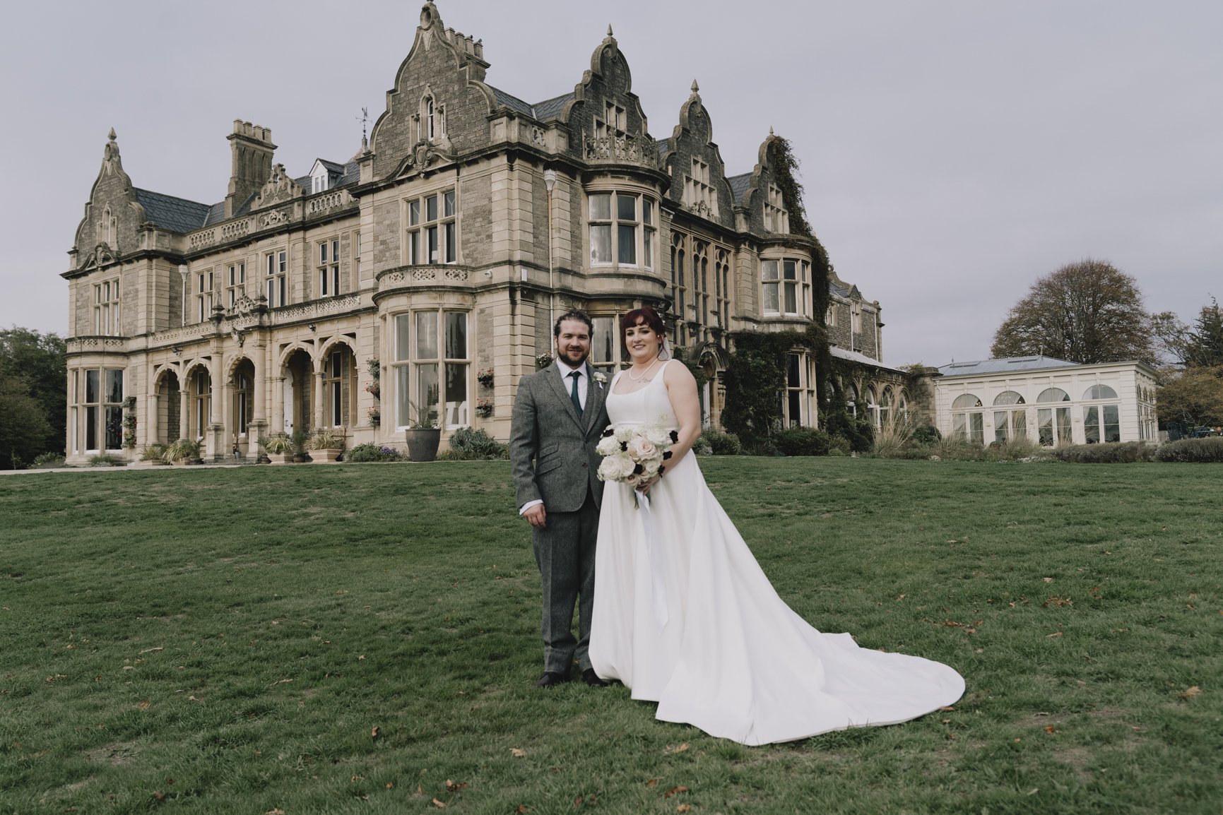 A bride and groom standing on grass in front of large historic mansion, Clevedon Hall, with the bride holding a bouquet of white flowers and both dressed in wedding attire .