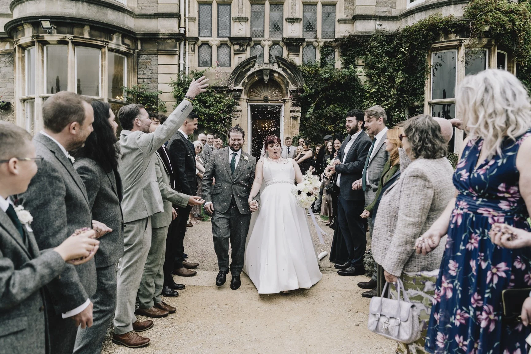 A newlywed couple walks hand in hand through a crowd outside a historic building during a wedding celebration, with guests throwing confetti in the air.