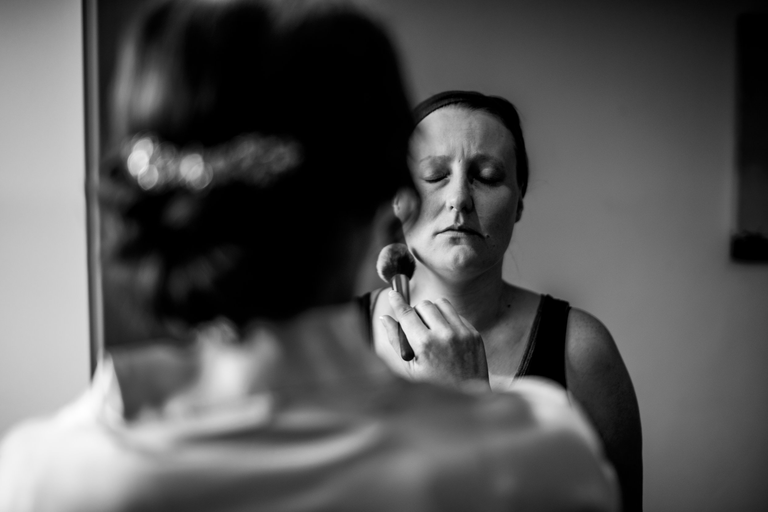 A woman is applying makeup with a brush while looking in a mirror, with her reflection visible during bridal prep. The image is in black and white.