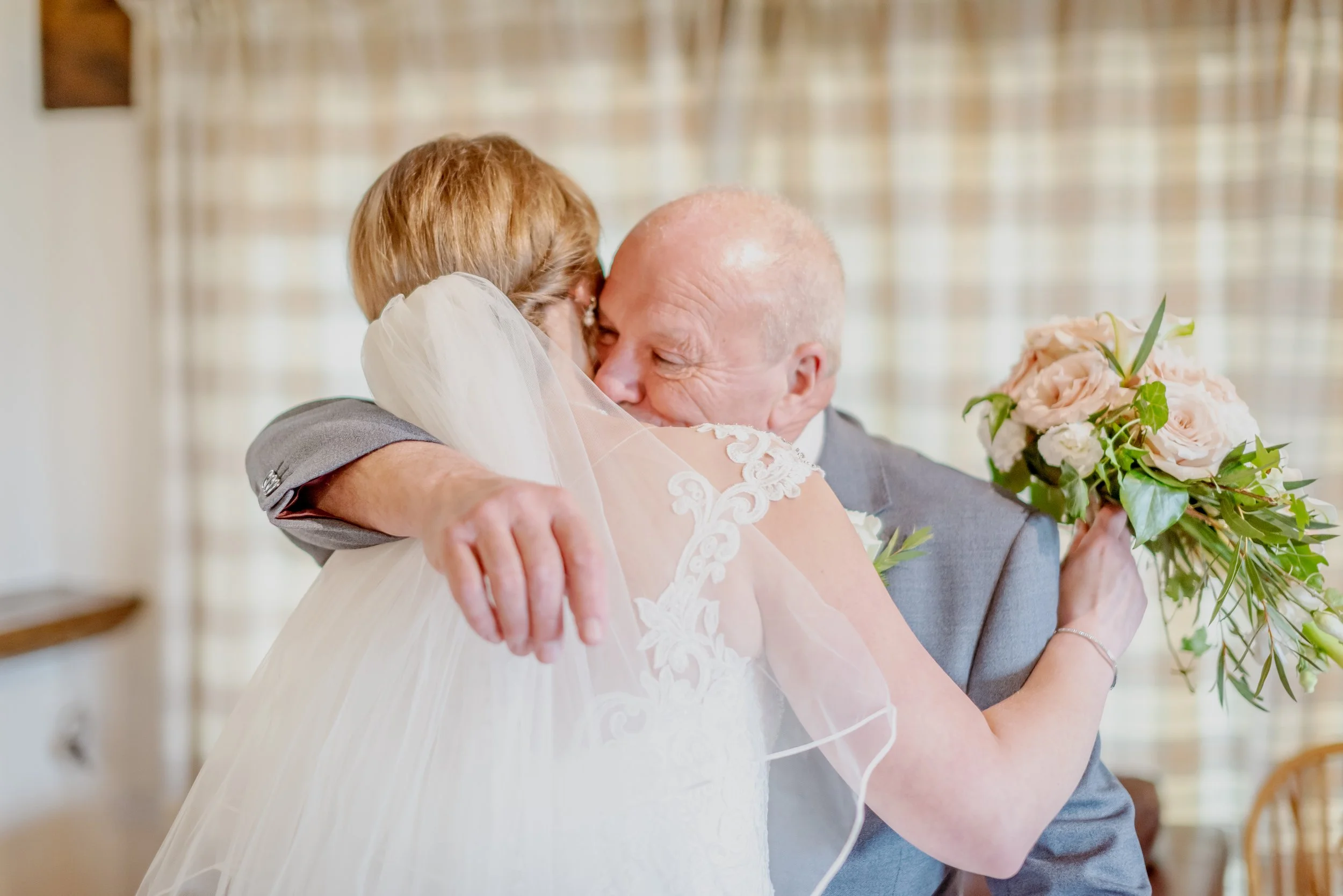 A bride and an older man, likely her father, sharing a hug and emotional moment at a wedding. The bride is holding a bouquet of pink roses and greenery.