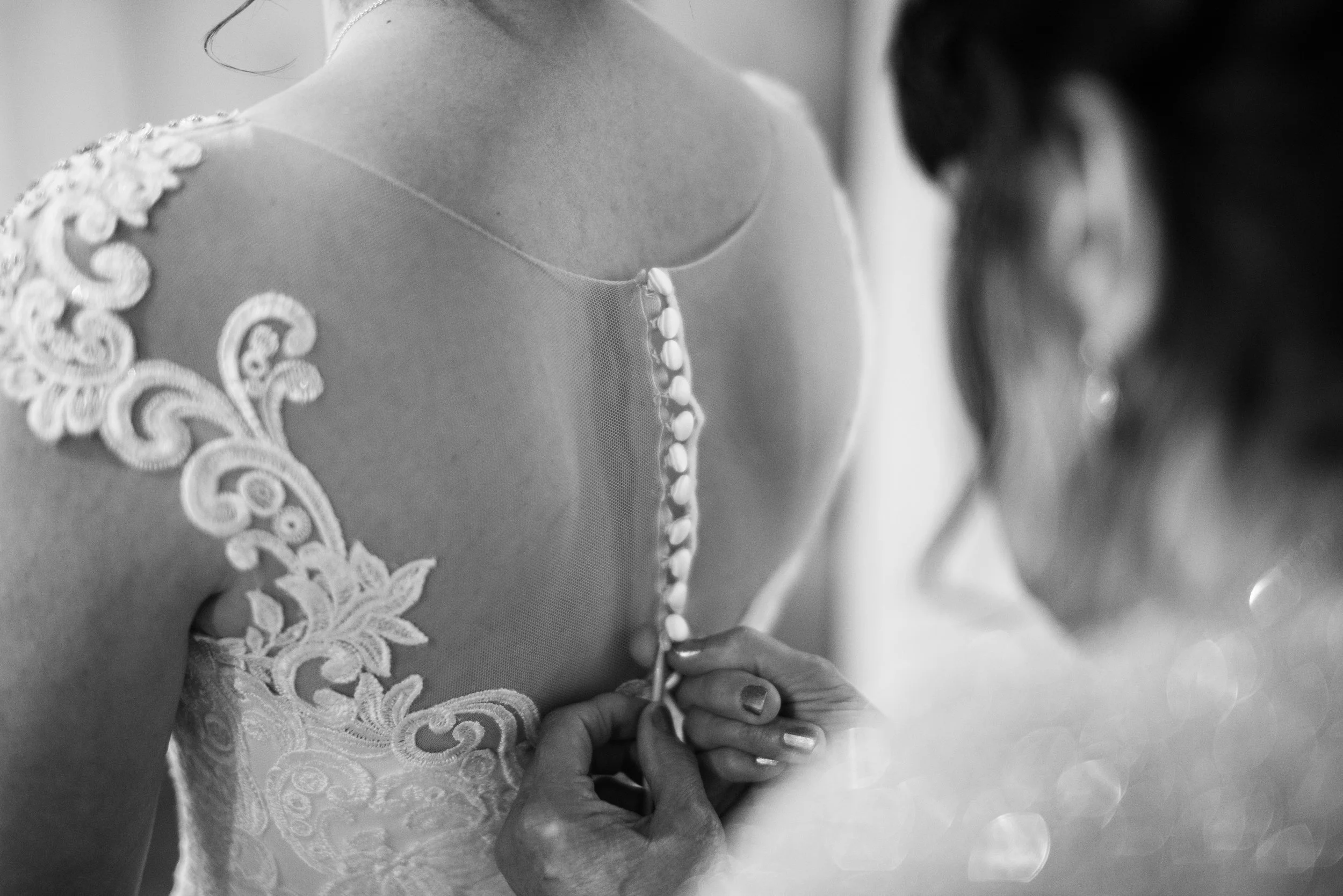 A woman in a wedding dress is buttoning up the back of her gown, with lace embroidery on the shoulder and upper back.
