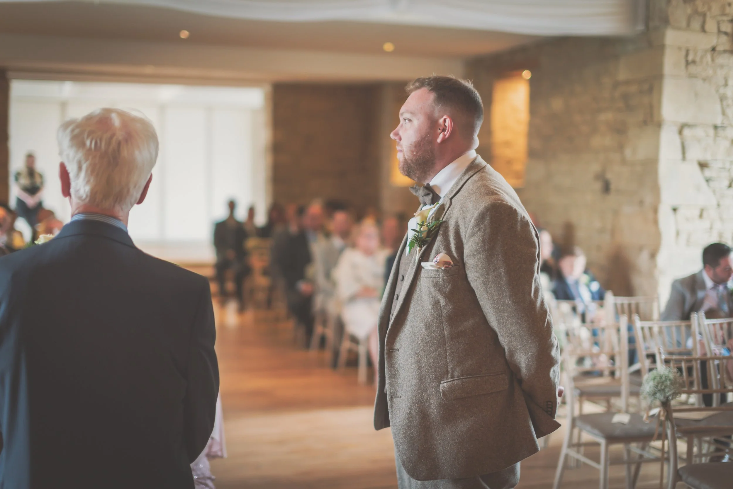 A groom in a brown suit with a boutonniere stands solemnly during a wedding ceremony, facing an officiant with his back to the camera at The Great Tythe Barn.