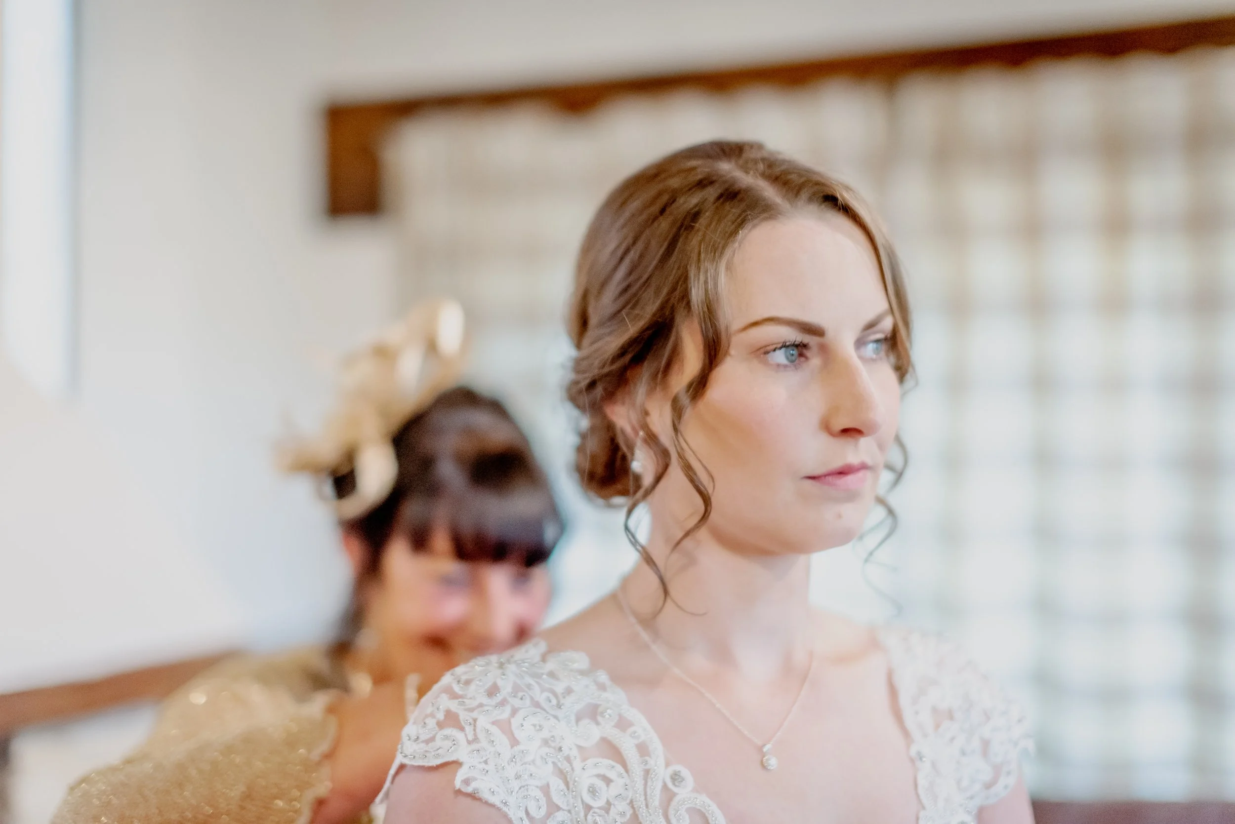 A bride with brown curly hair in a white lace dress and pearl necklace preparing for her wedding, with another woman in a gold dress assisting her taken by Cotswold Wedding Photographer.