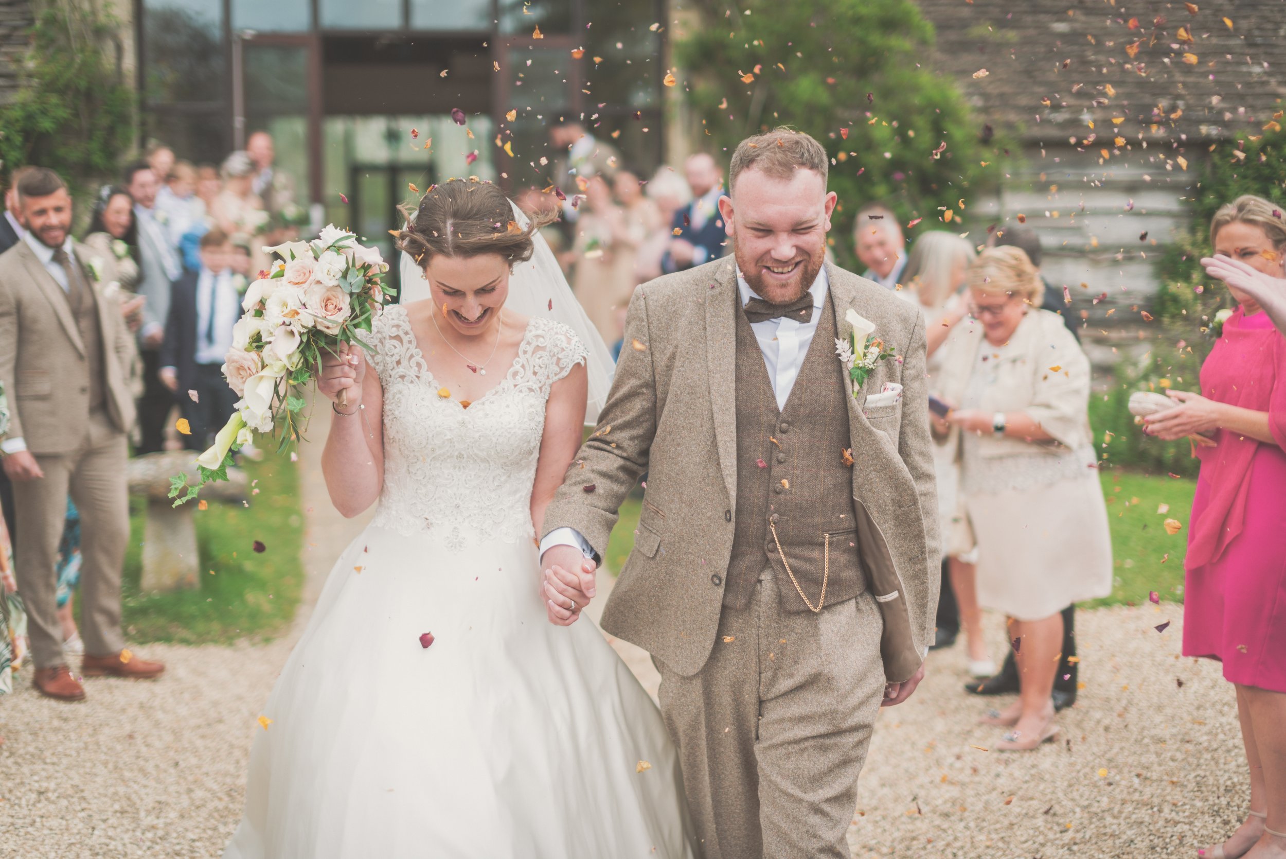 A bride and groom walking hand in hand after wedding, surrounded by guests throwing confetti, outdoors in a garden setting taken by Cotswold Wedding Photographer