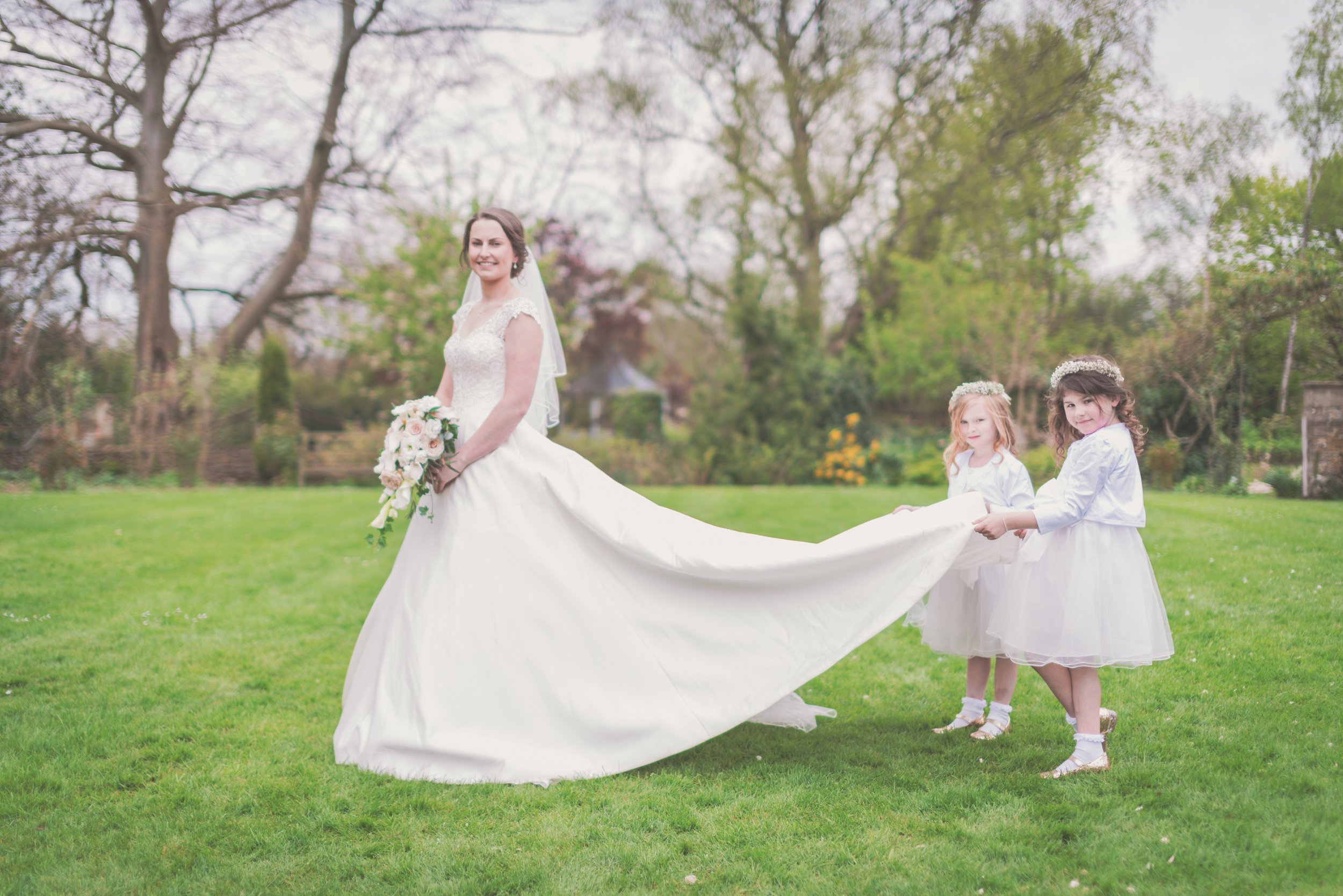 Bride in a white wedding dress holding a bouquet of flowers, standing on a grassy field, with two flower girls in white dresses holding the train of her dress, in a park-like setting with trees and bushes in the background taken at Great Tythe Barn