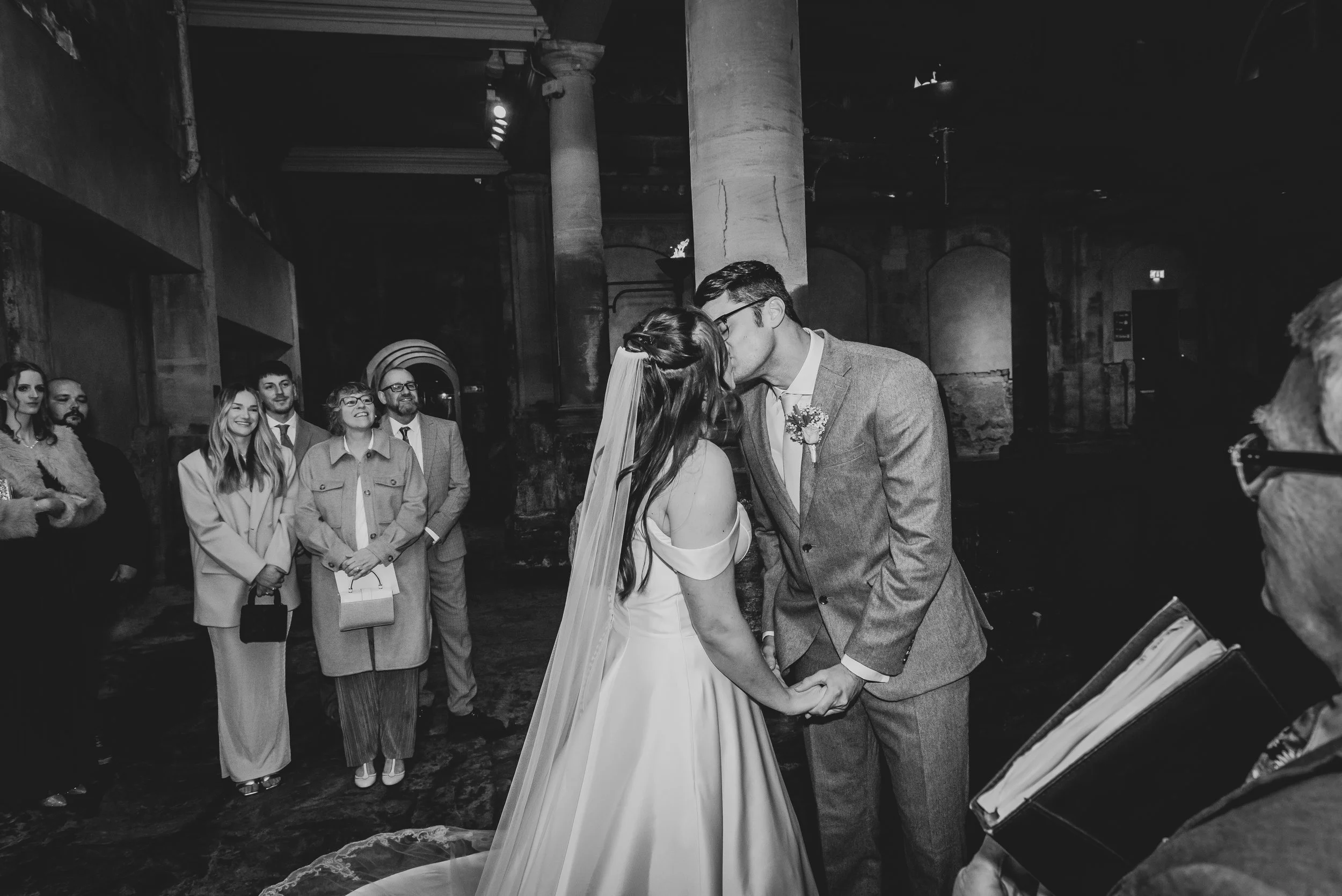 Bride and groom sharing their first kiss as newlyweds beside the Great Bath at Roman Baths in Bath