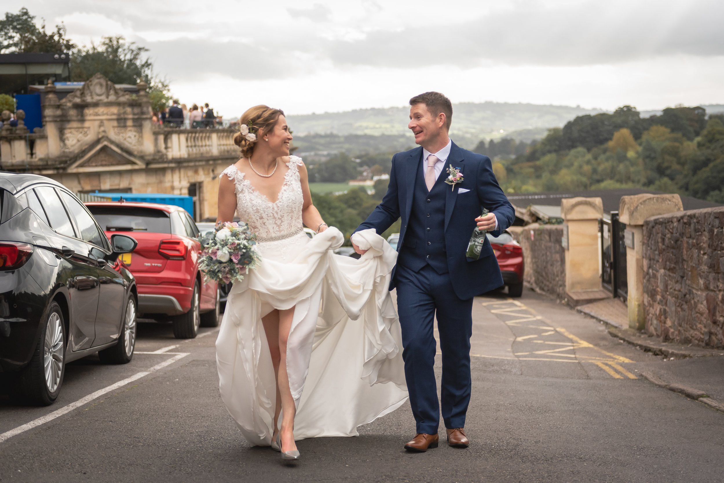 Bride and groom walking together outdoors, the bride lifting her wedding dress slightly, smiling at each other, the groom holding a bottle, both dressed in wedding attire, with cars parked along the street and scenic landscape in Bristol.