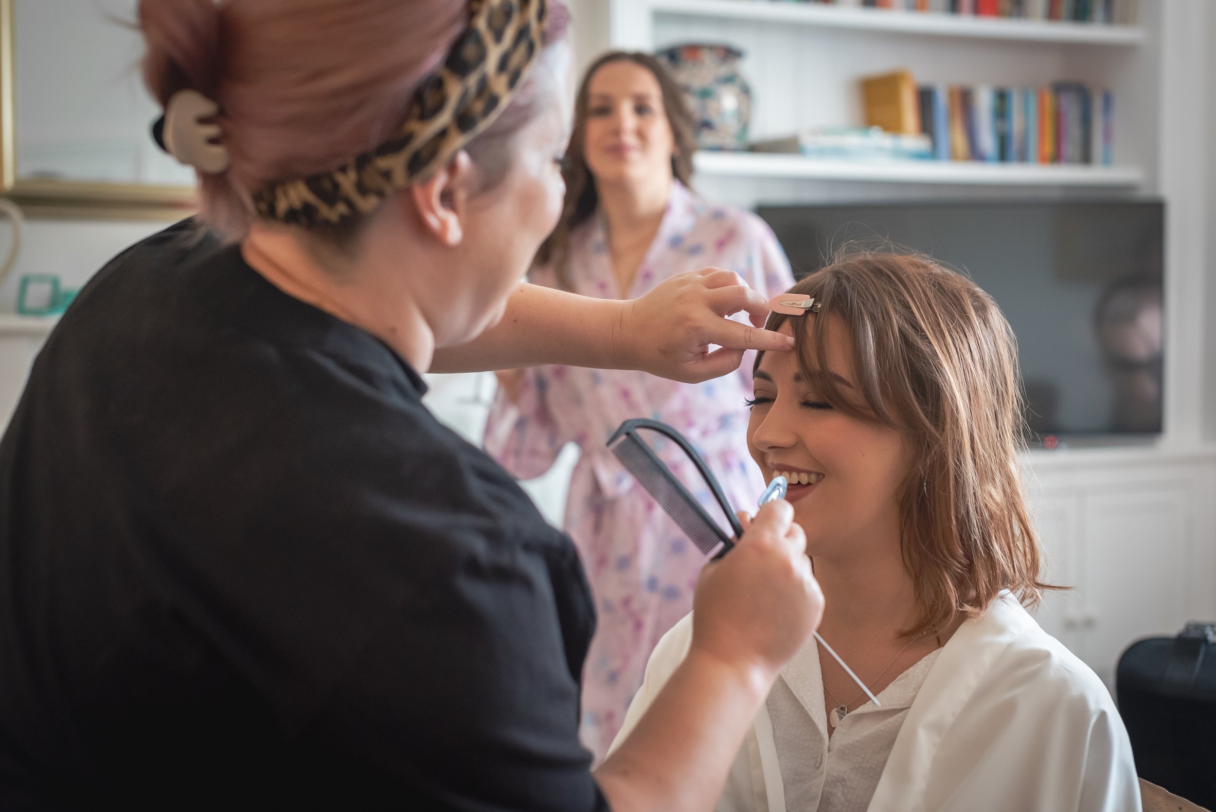 Bride is getting her makeup done by a makeup artist while a person in the background looks on, all in a bright room with books on shelves in Devon.