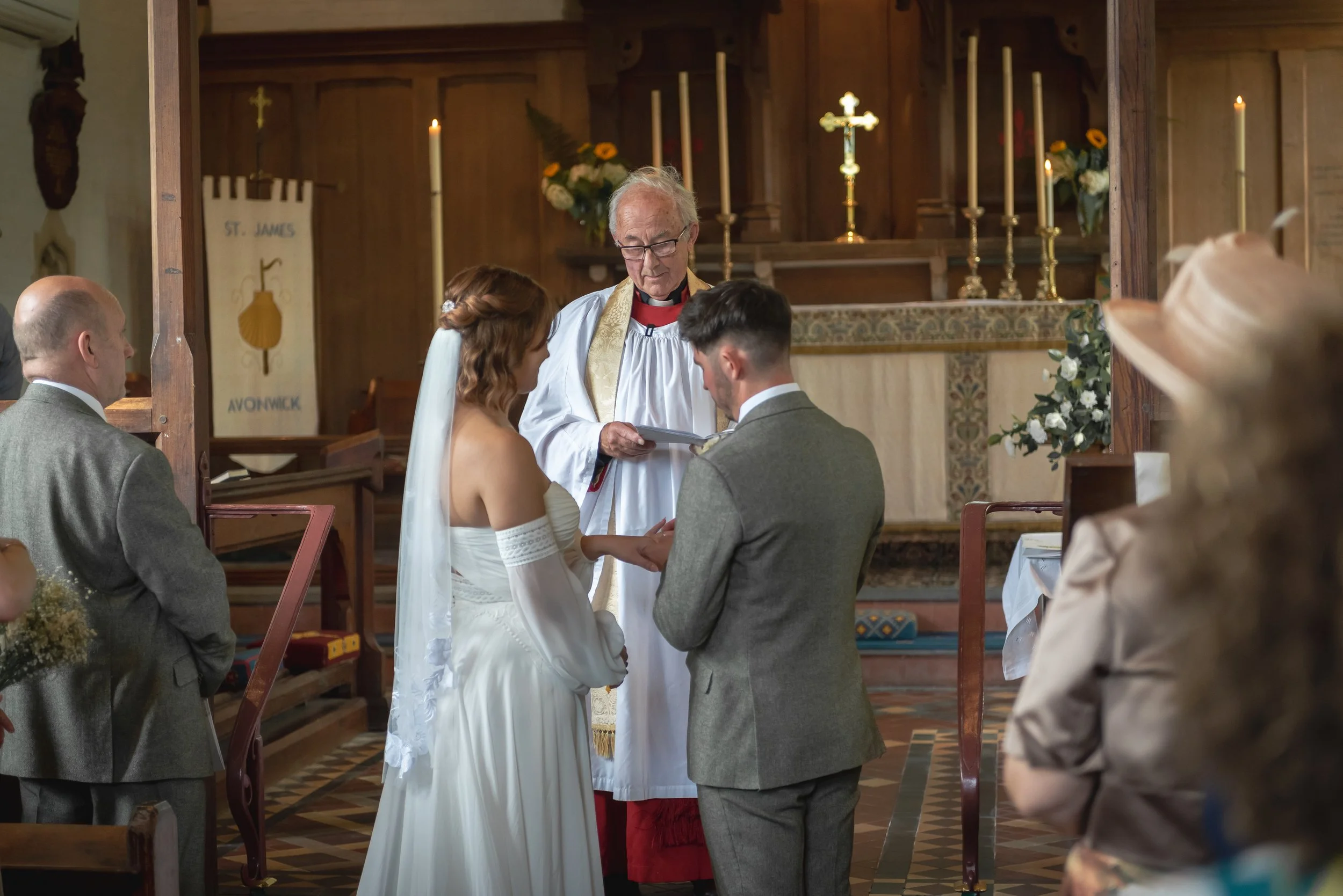 A wedding ceremony taking place inside a church with the bride and groom holding hands in front of a pastor, during the vows. The bride is wearing a white dress with an off-the-shoulder design and veil, and the groom is dressed in a gray suit. There are people seated watching, candles on the altar, and floral arrangements.