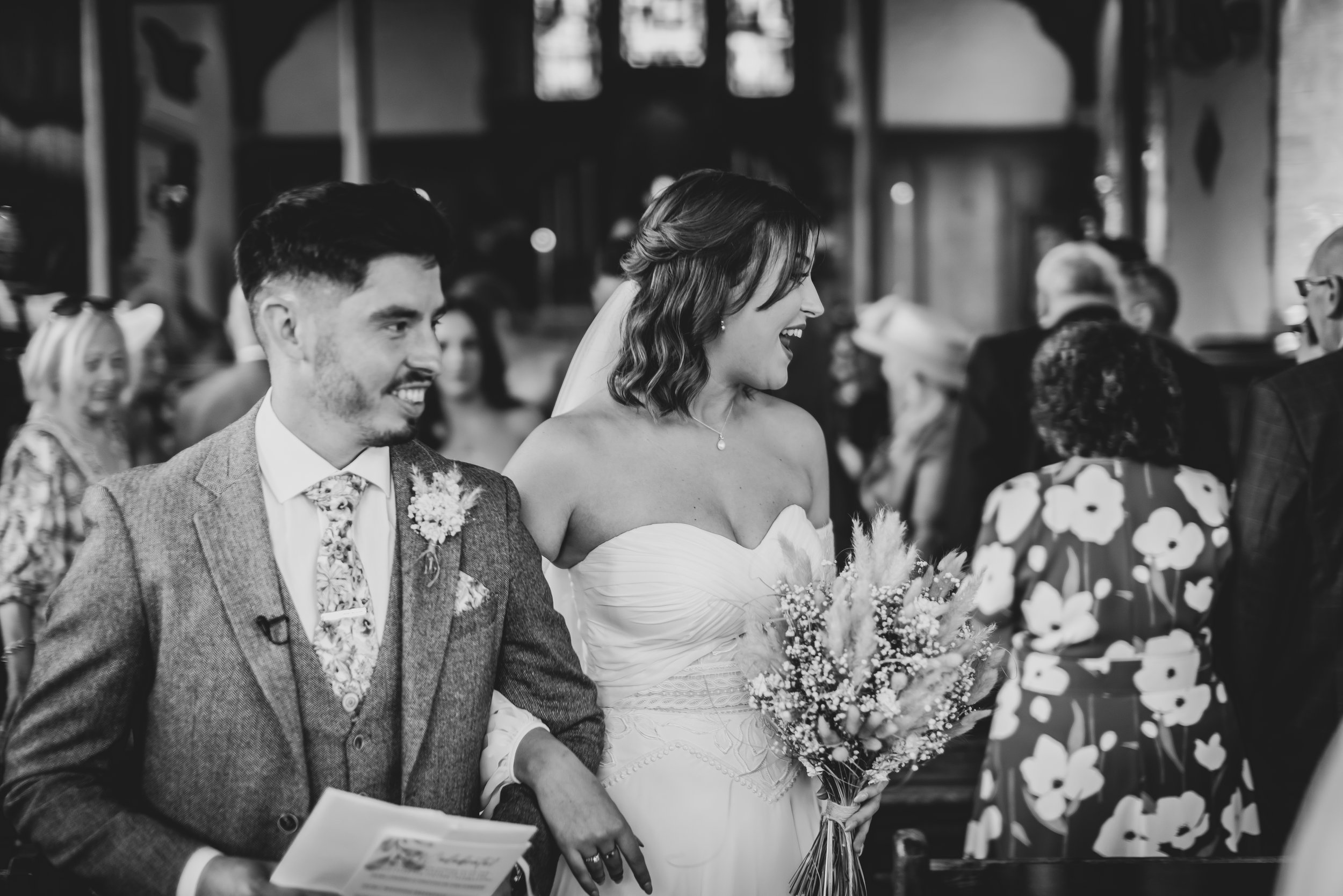 A couple at a wedding ceremony. The woman is wearing a strapless wedding dress and holding a bouquet, smiling and looking to her left. The man is dressed in a suit with a floral tie and a boutonniere, smiling and holding papers in his hand. They are surrounded by guests.