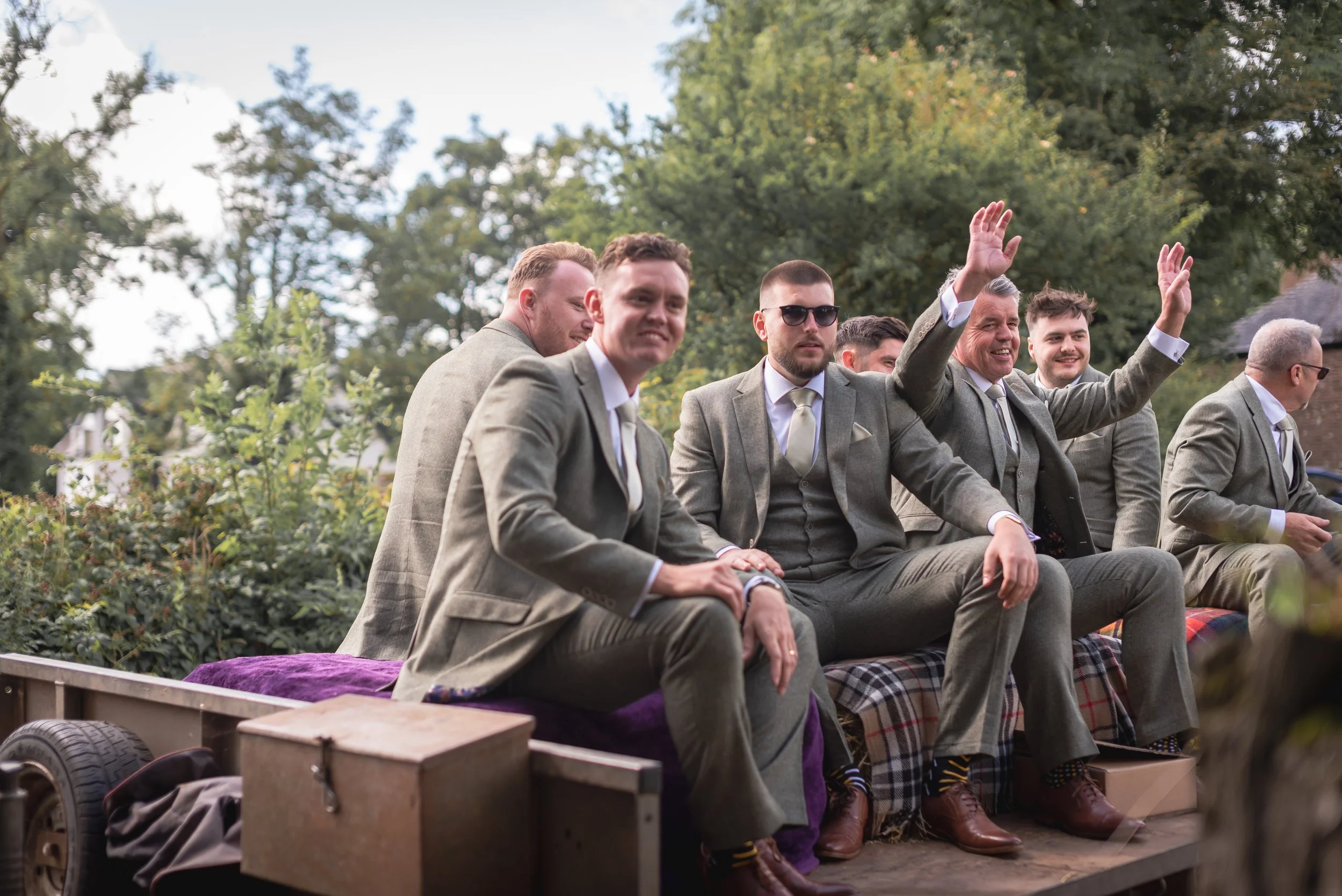 A group of men dressed in gray suits sitting on a decorated float during an outdoor event, with some waving and smiling, surrounded by green trees.