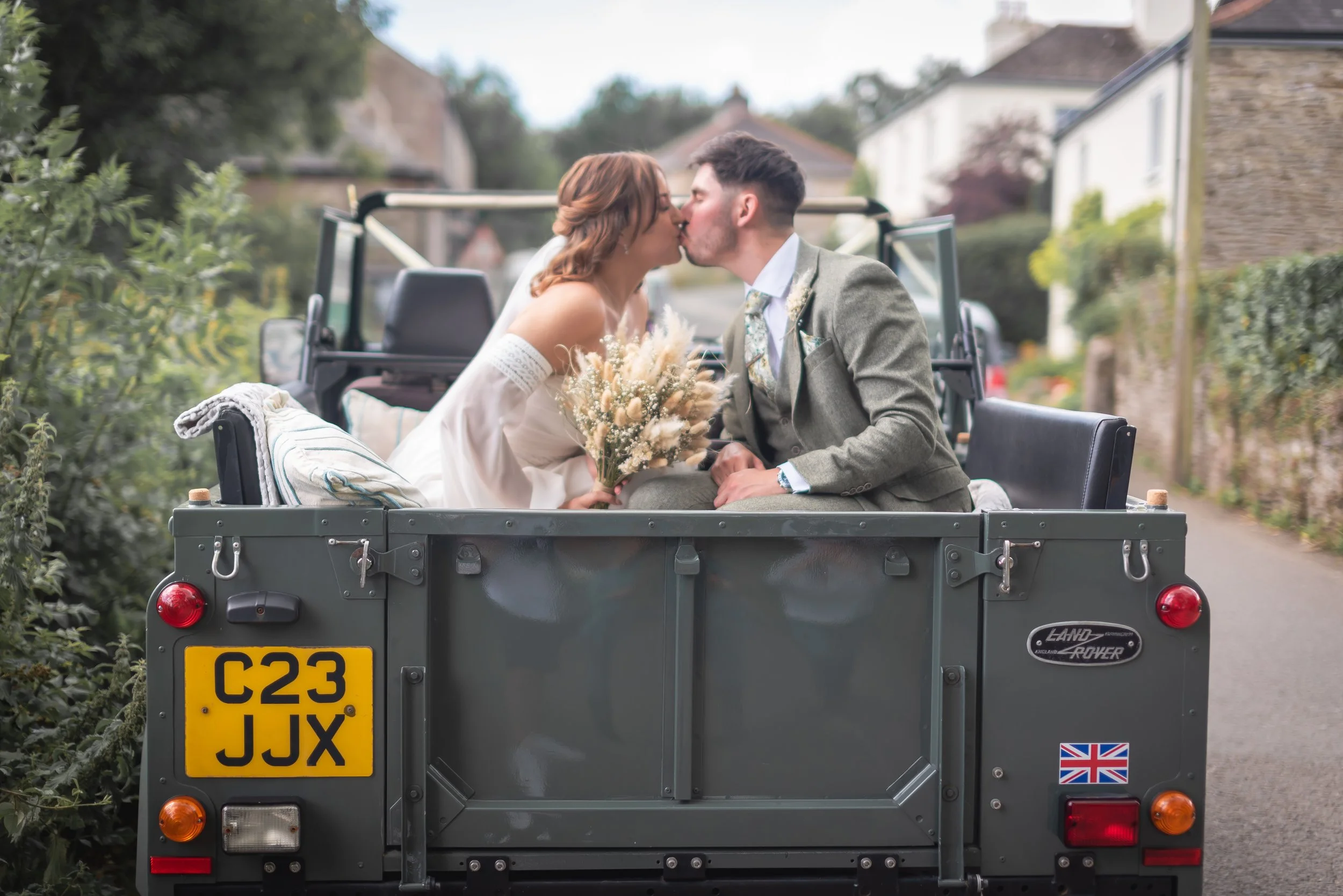 A newlywed couple sharing a kiss in the back of a vintage Land Rover on a country road, with houses and trees in the background captured by Devon Wedding Photographer