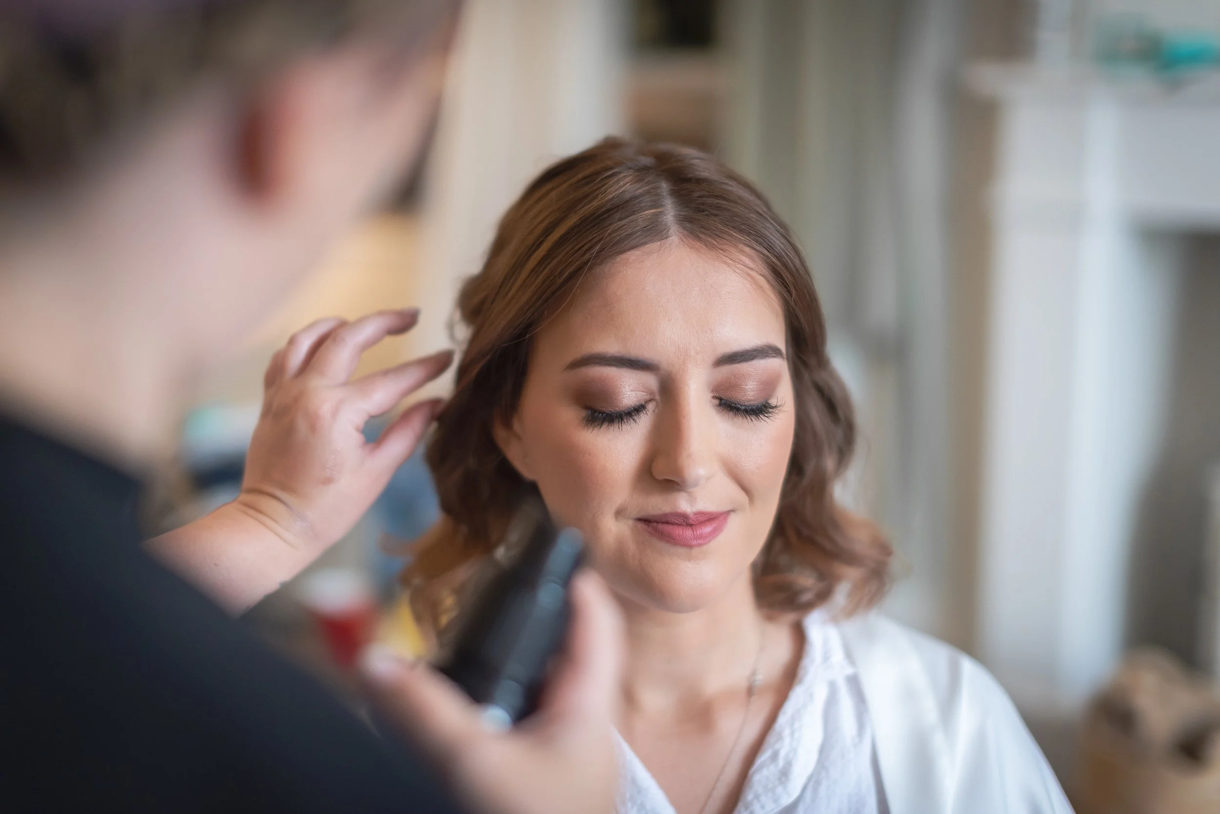 A woman with closed eyes getting her makeup done by a makeup artist holding a makeup brush ready for her wedding in devon.