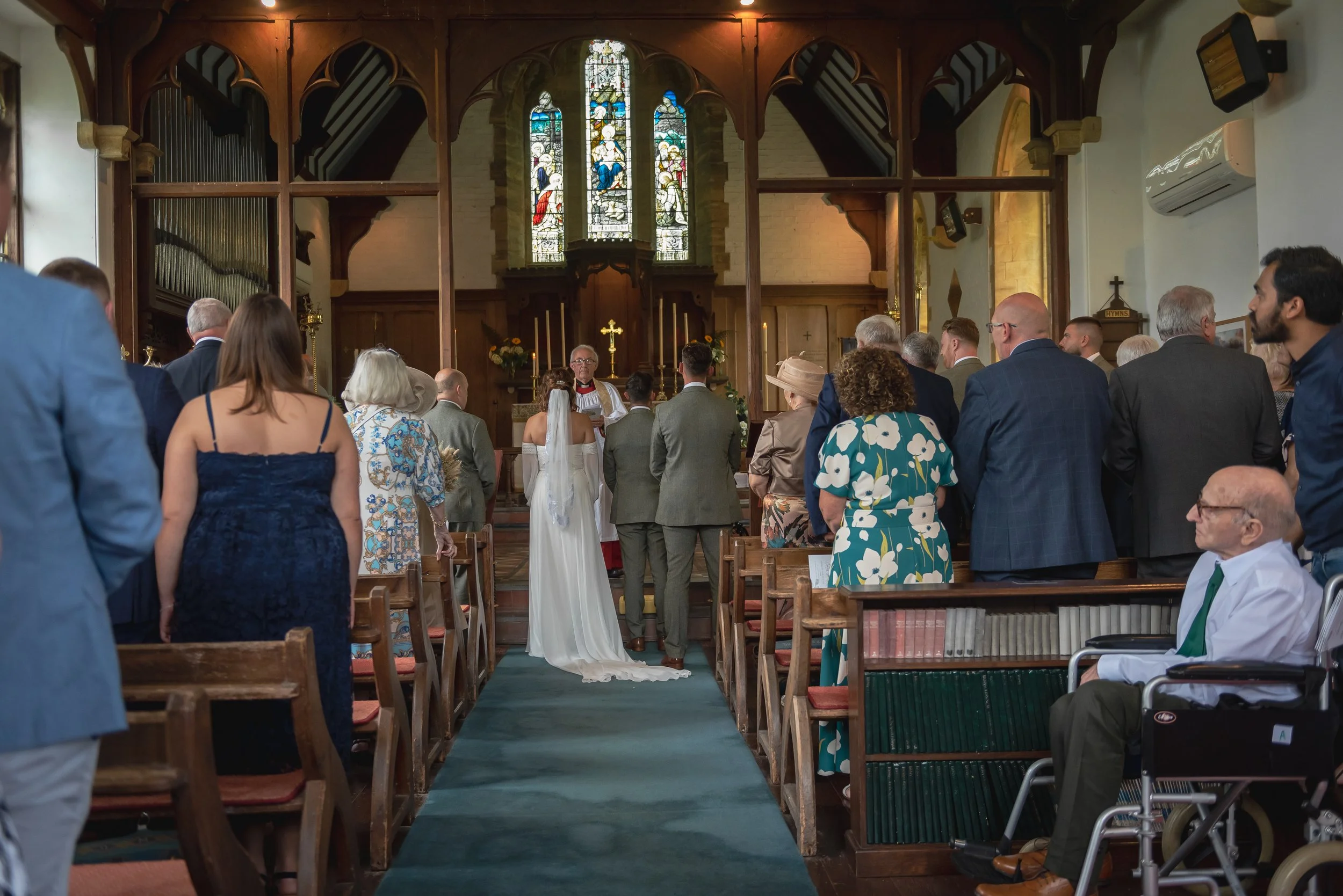 A wedding ceremony inside a church with the bride and groom standing at the altar with the officiant, surrounded by guests seated in pews, stained glass windows above, and religious symbols on the altar at St James Church in Torquay.