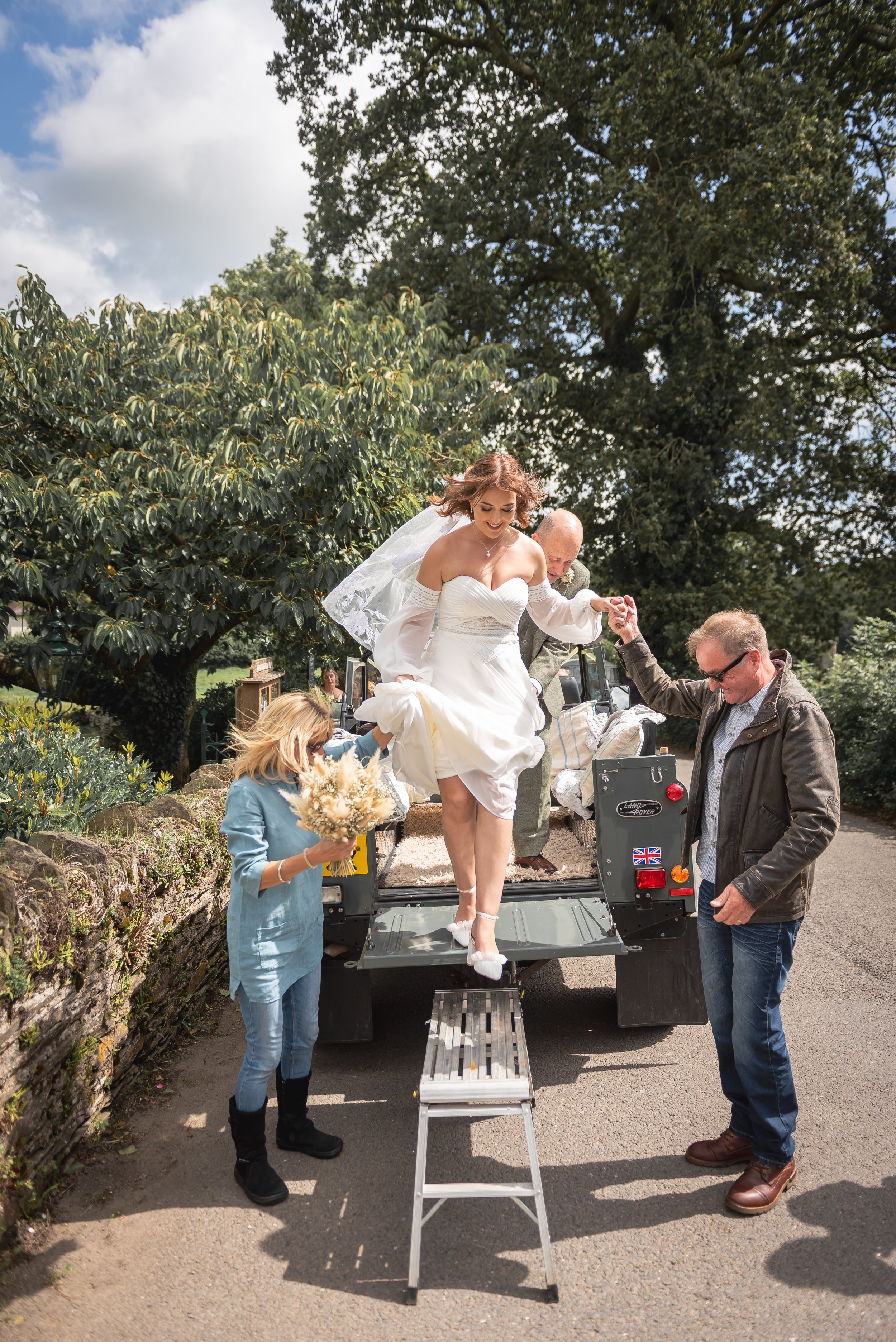 A bride in a white wedding dress stepping down from a vehicle, assisted by two men. A woman holding a bouquet of flowers stands nearby. The scene is outdoors with trees and a cloudy sky during wedding near Devon.