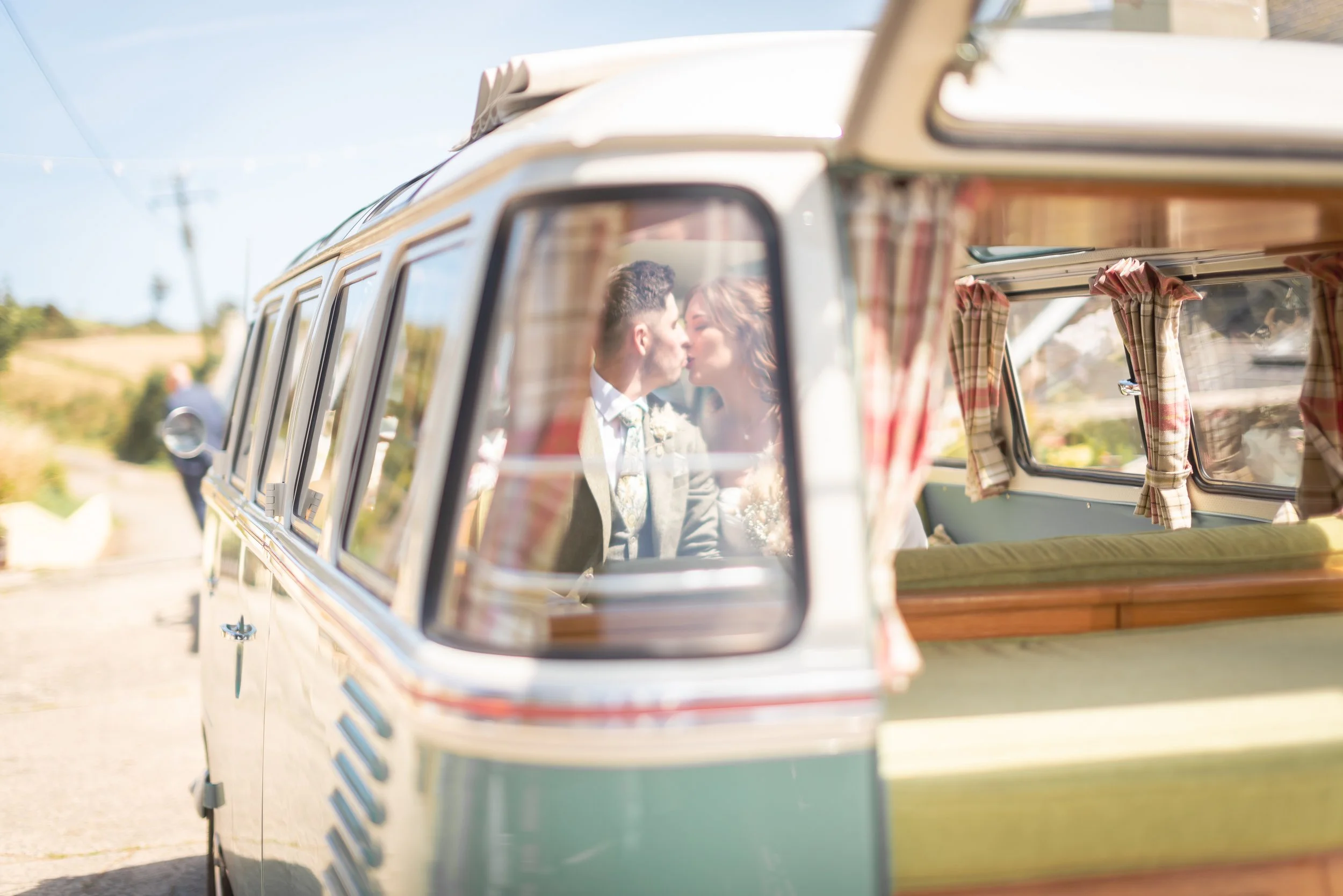 A couple is kissing inside a vintage camper van with checkered curtains, viewed through a side window, on a sunny day during Devon Wedding Photography.