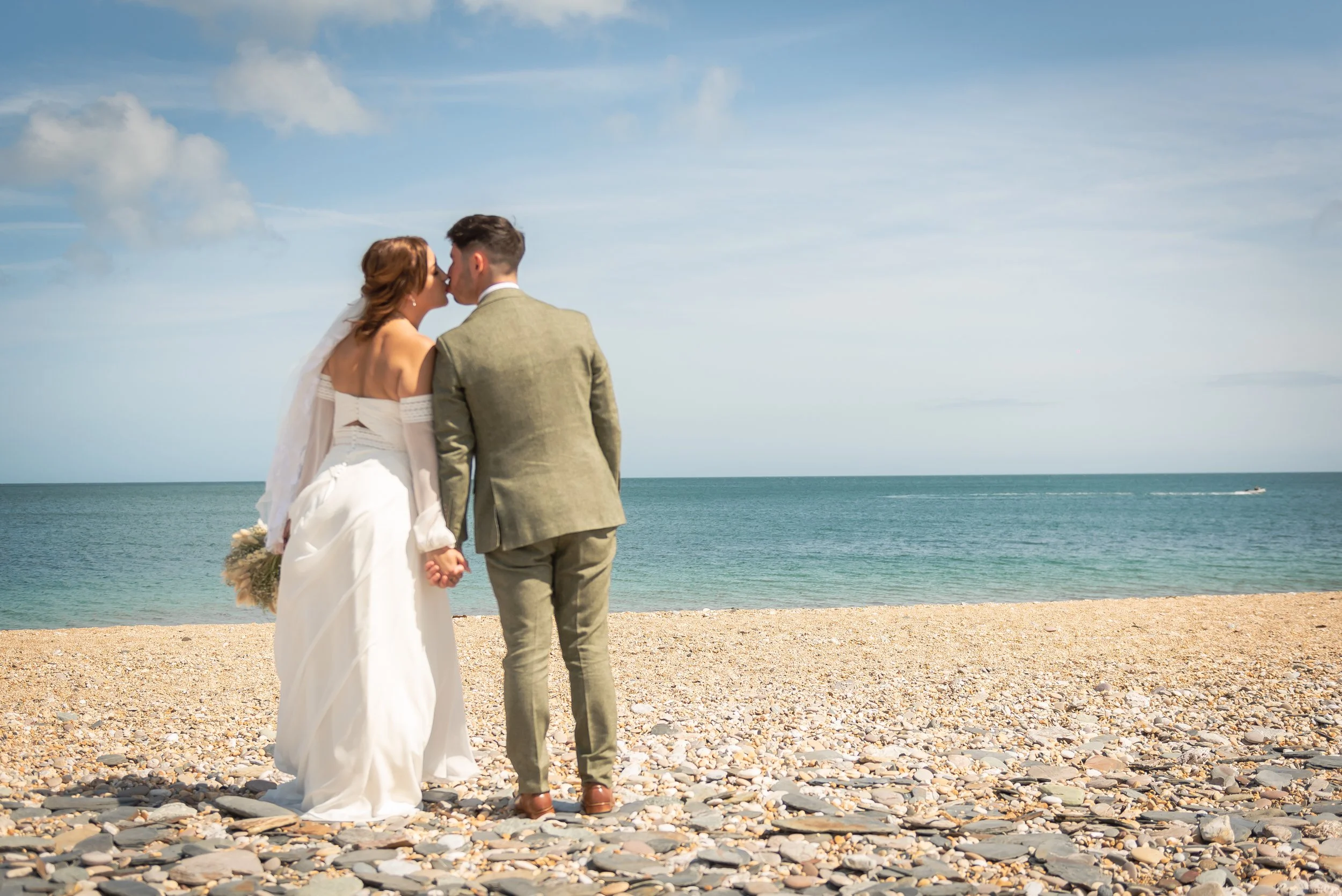 A bride and groom kissing on a pebble beach with the ocean and blue sky in the background capture by Devon Wedding Photographer.