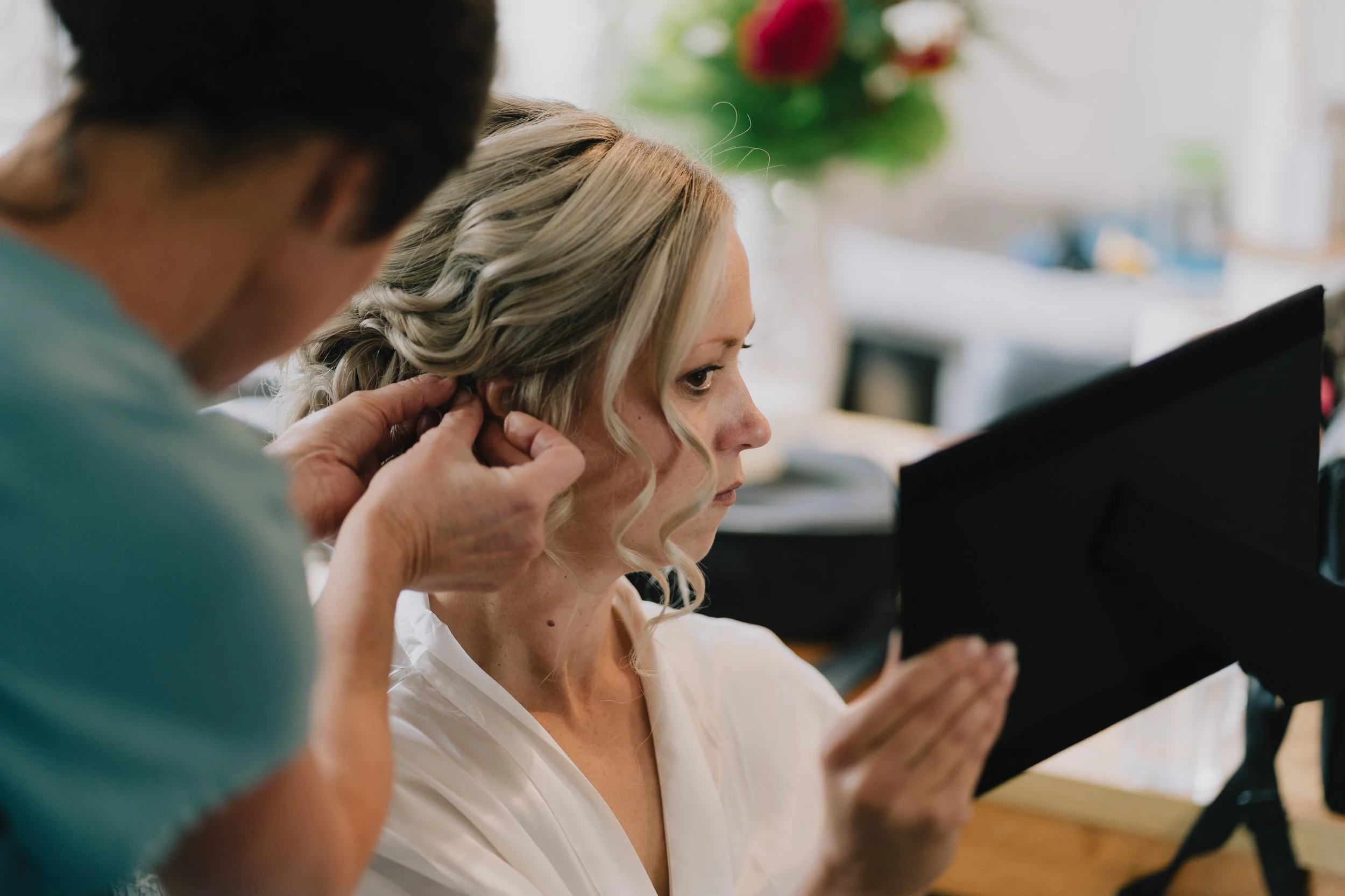 A woman with blonde hair receives hair styling from a person, while she looks at herself in a mirror.