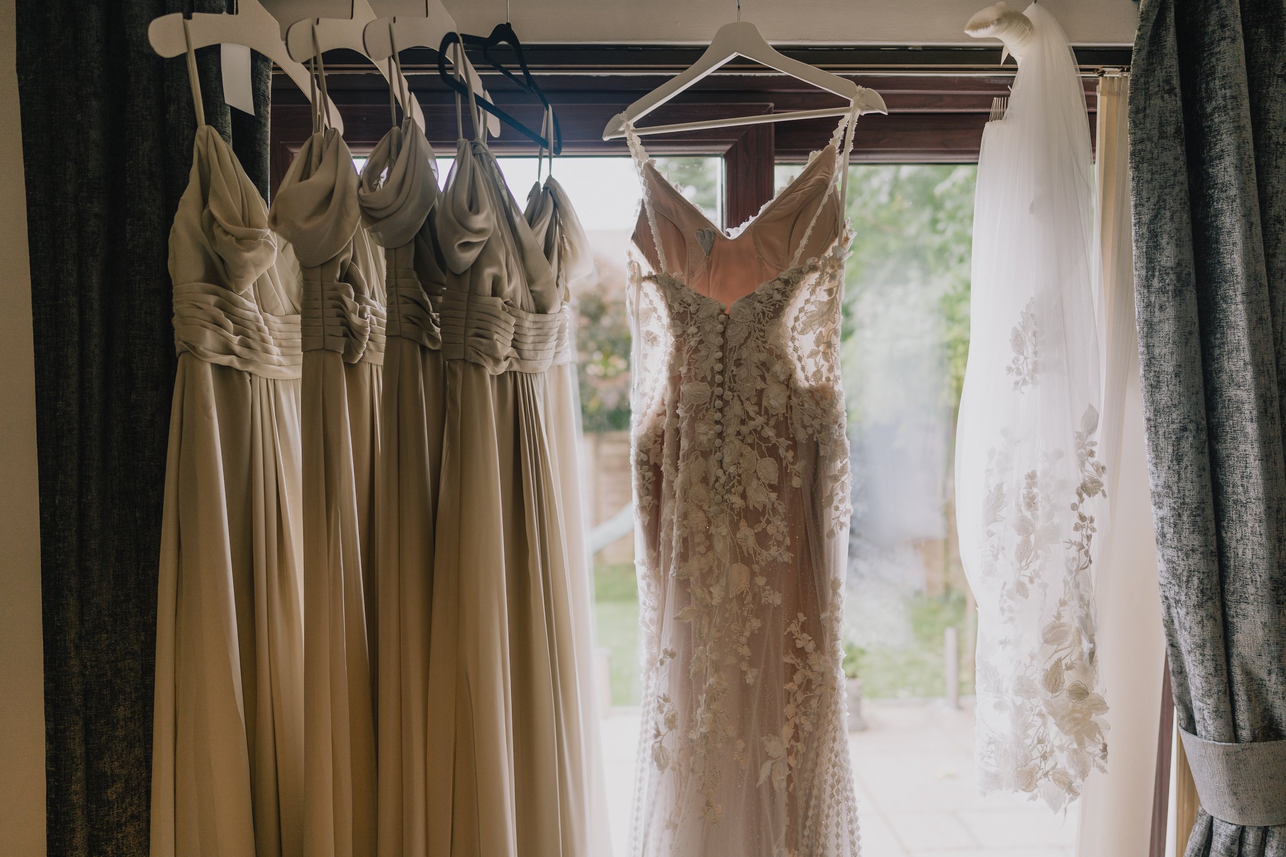 A collection of wedding dresses hanging on a window curtain rod, including a lace wedding gown and a strapless dress, with natural light outdoors.