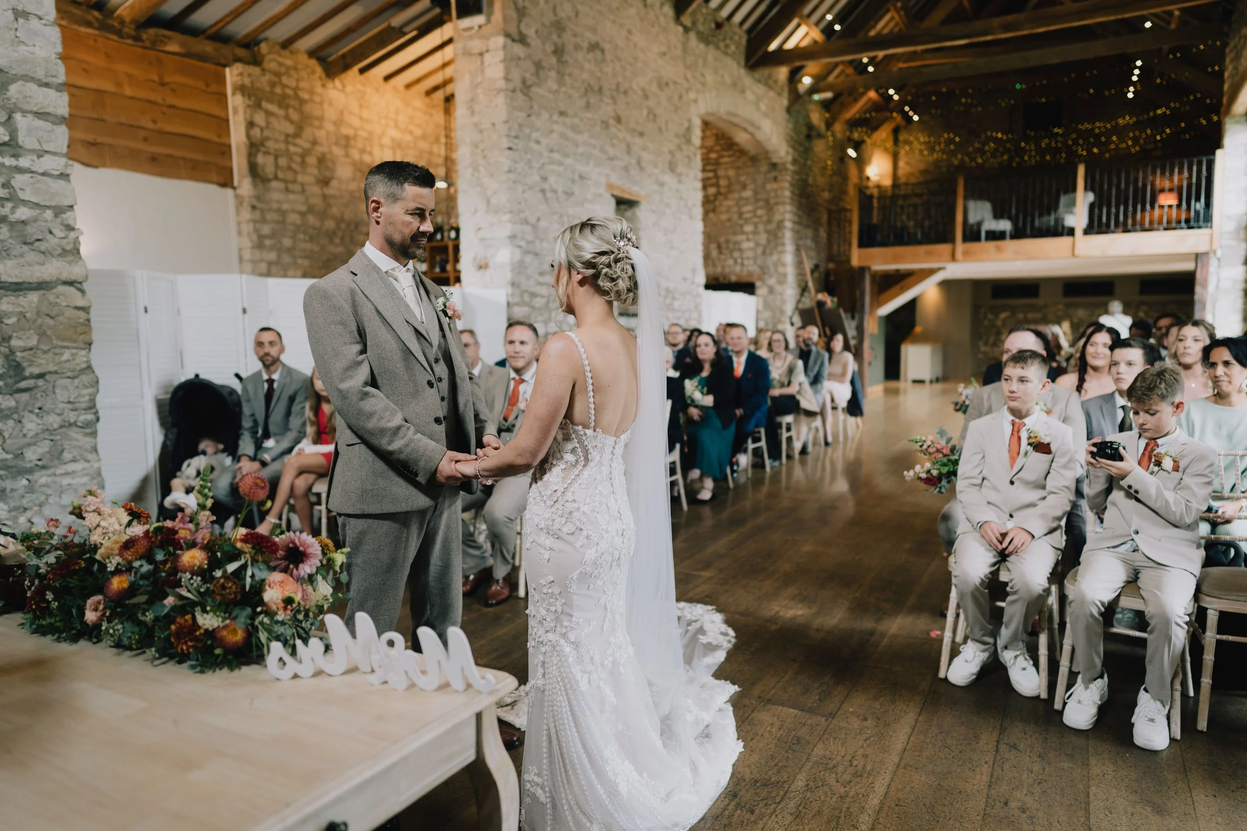 A wedding ceremony taking place indoors with a bride and groom holding hands and exchanging vows, surrounded by guests seated on chairs, in a rustic venue with stone walls and wooden beams.