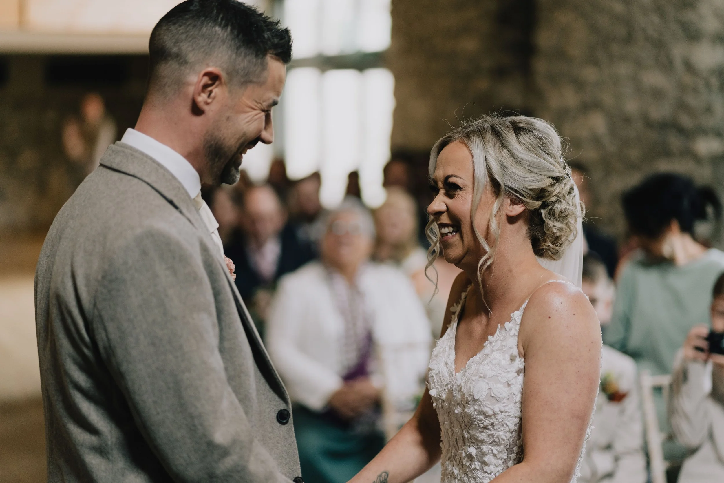 A bride and groom sharing a joyful moment during their wedding ceremony, holding hands and smiling at each other.