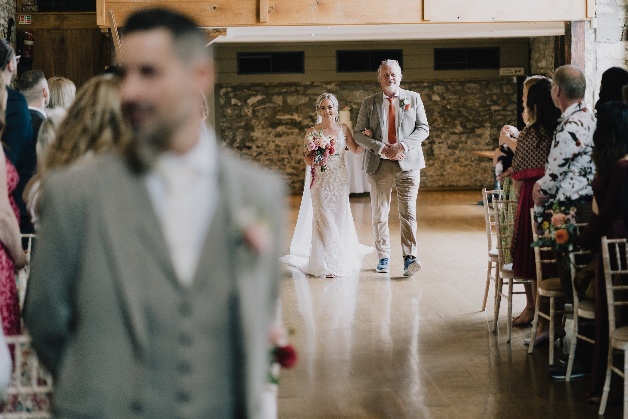 Bride walking down the aisle arm-in-arm with a man in a light gray suit during a wedding ceremony in a rustic venue with stone walls and wooden decorations.