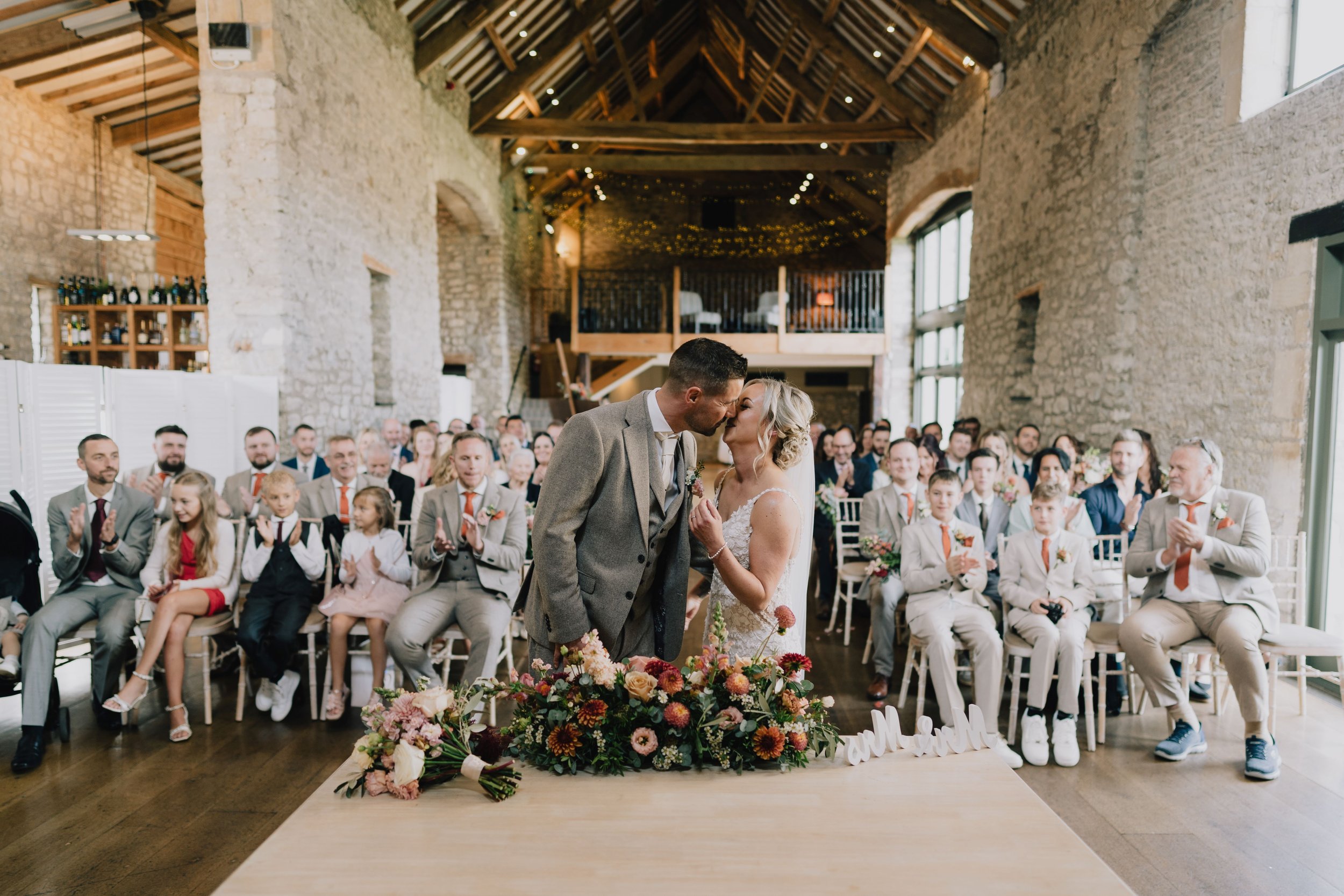 A bride and groom sharing a kiss at their wedding ceremony, surrounded by seated guests in a rustic stone venue with large windows and wooden beams.