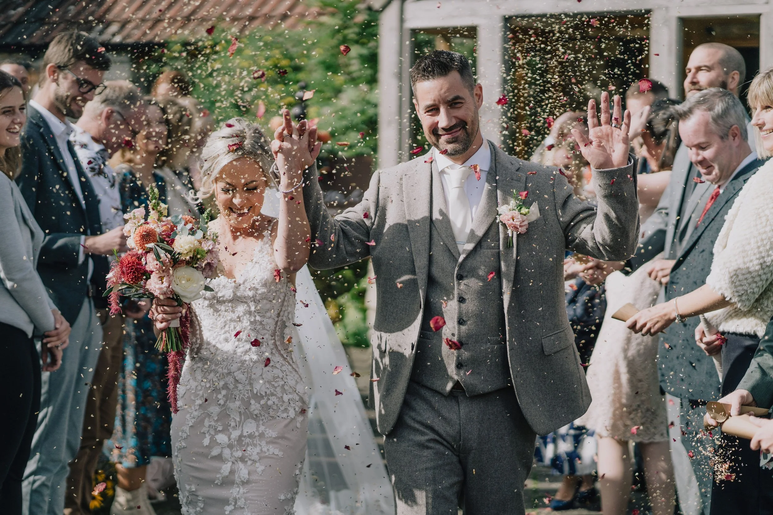 A newlywed couple walking through a shower of flower petals surrounded by wedding guests outdoors.