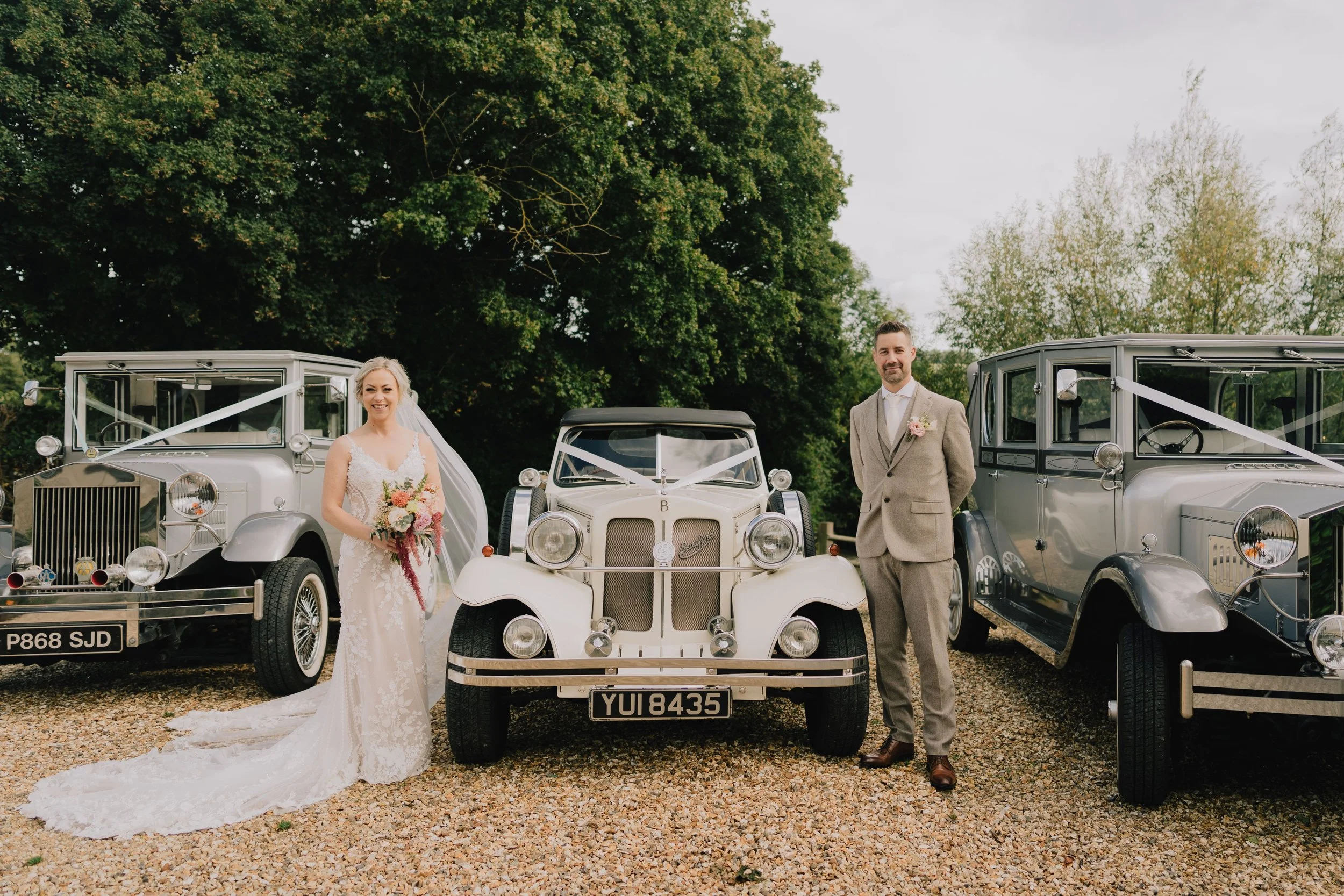 Bride in a white lace wedding dress and veil holding a bouquet, standing next to a groom in a beige suit, both smiling, with vintage cars and lush trees in the background.